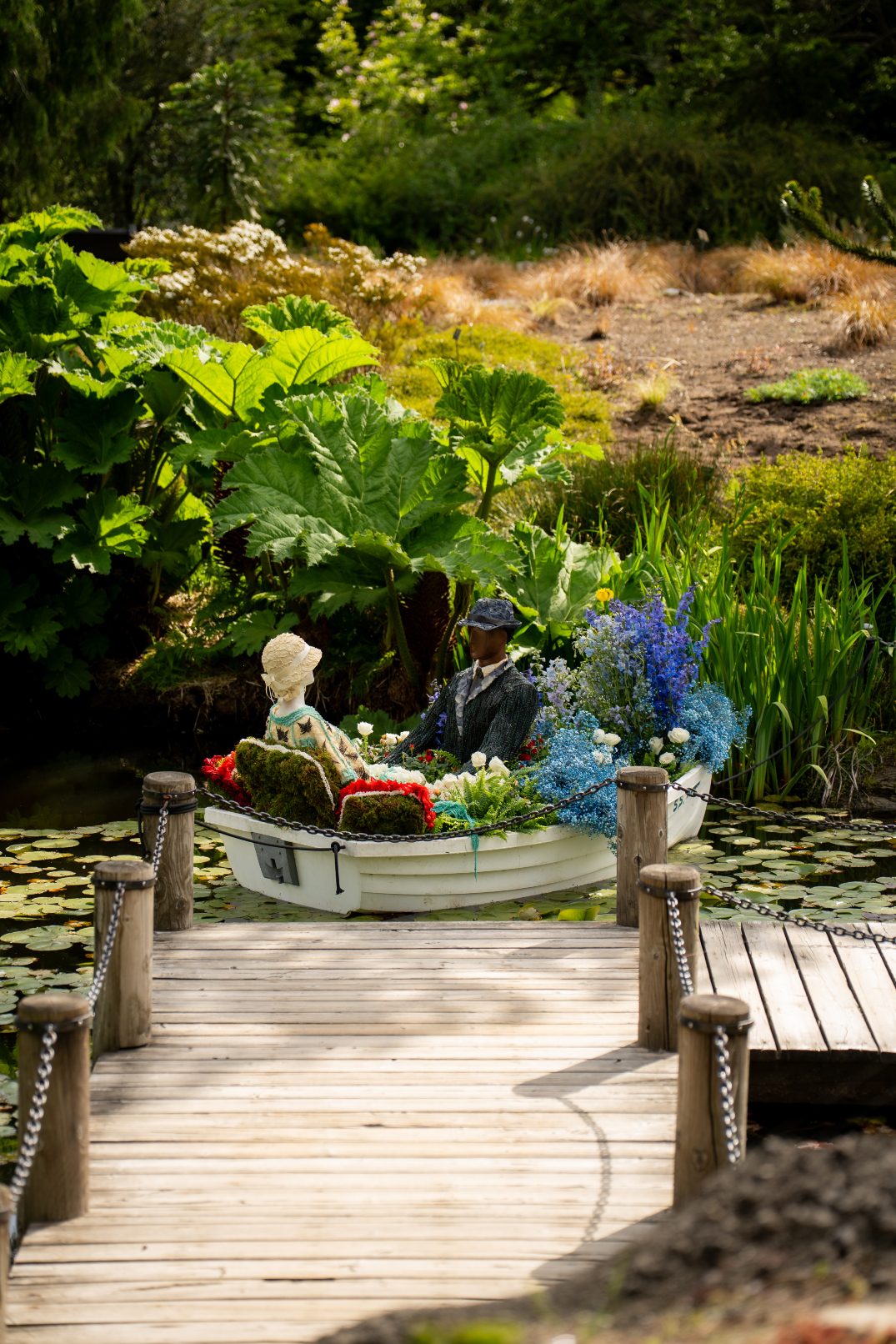 A small white rowboat adorned with vibrant blue, white and red flowers sits near a dock surrounded by lily pads at VanDusen Botanical Gardens.