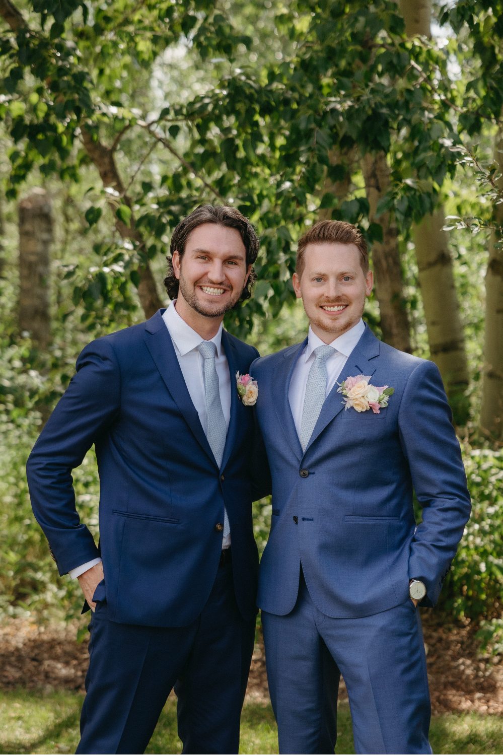 The groom poses with one of his groomsmen, both smiling broadly in sharp blue suits and pastel boutonnieres, standing beneath the trees.