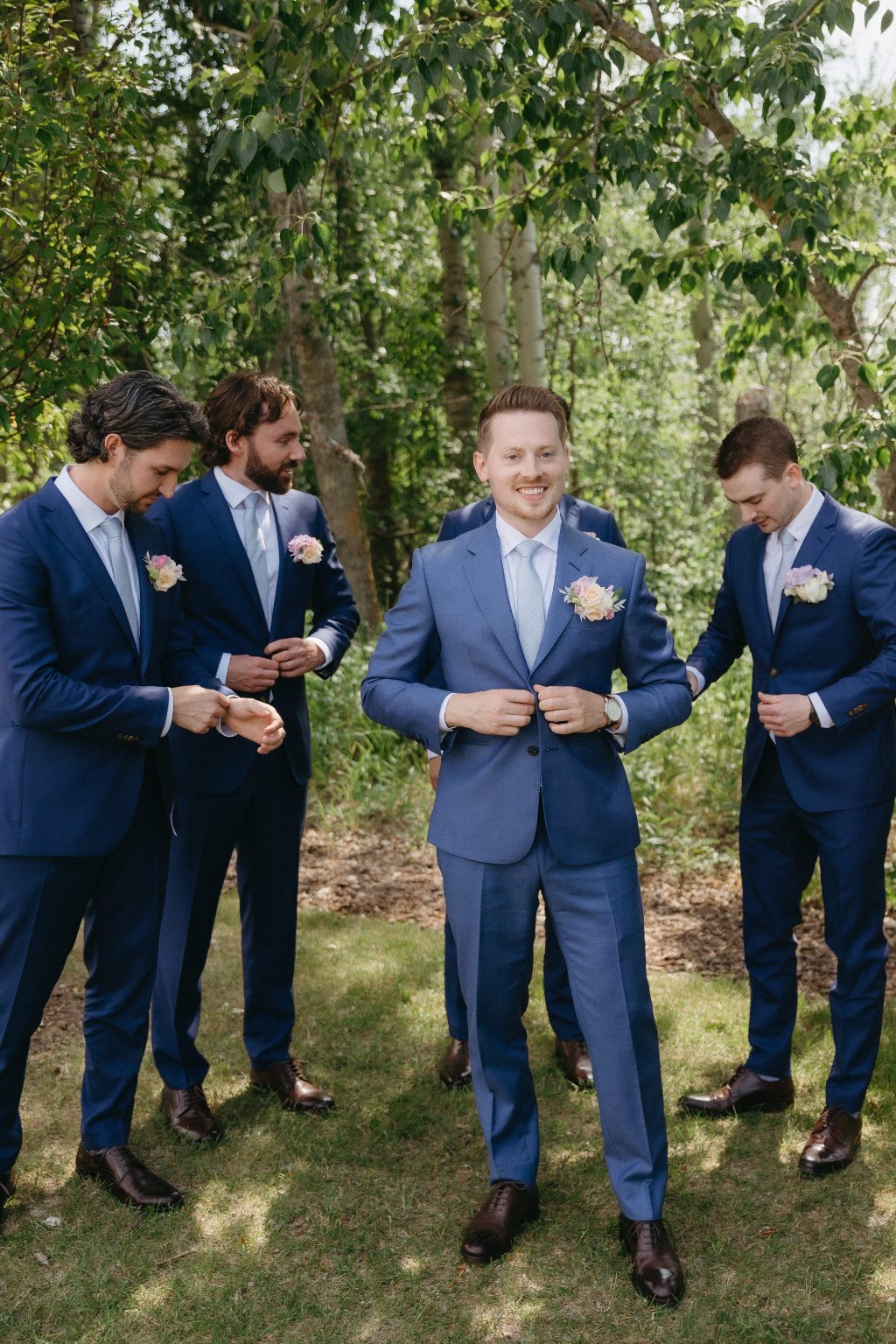 The groom stands centered and smiling as his groomsmen adjust their suits around him in a candid, lighthearted moment beneath the trees.
