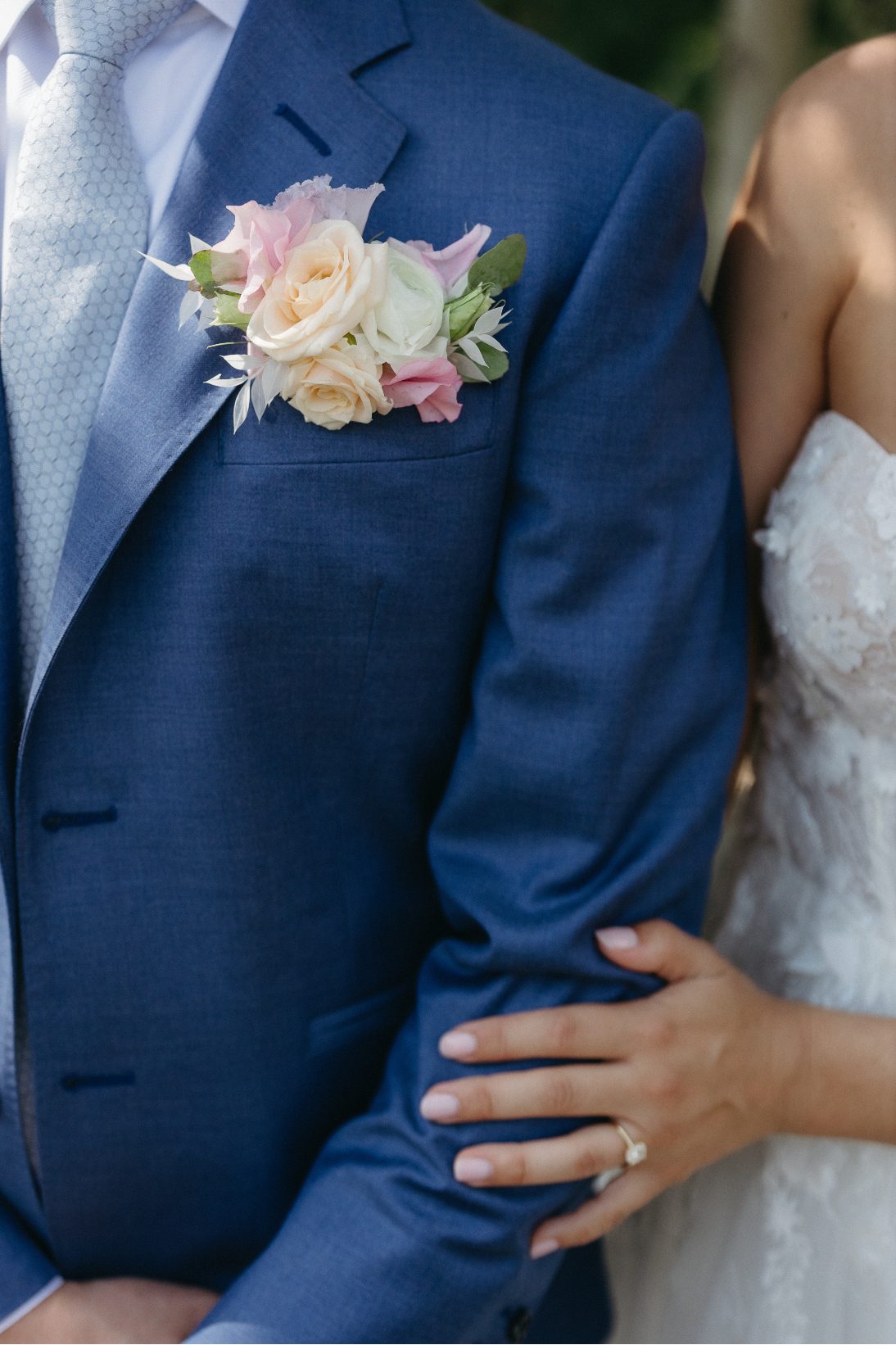 A close up of the groom's blue suit jacket and floral boutonniere, with the bride's hand gently resting on his arm, showcasing her stunning ring.