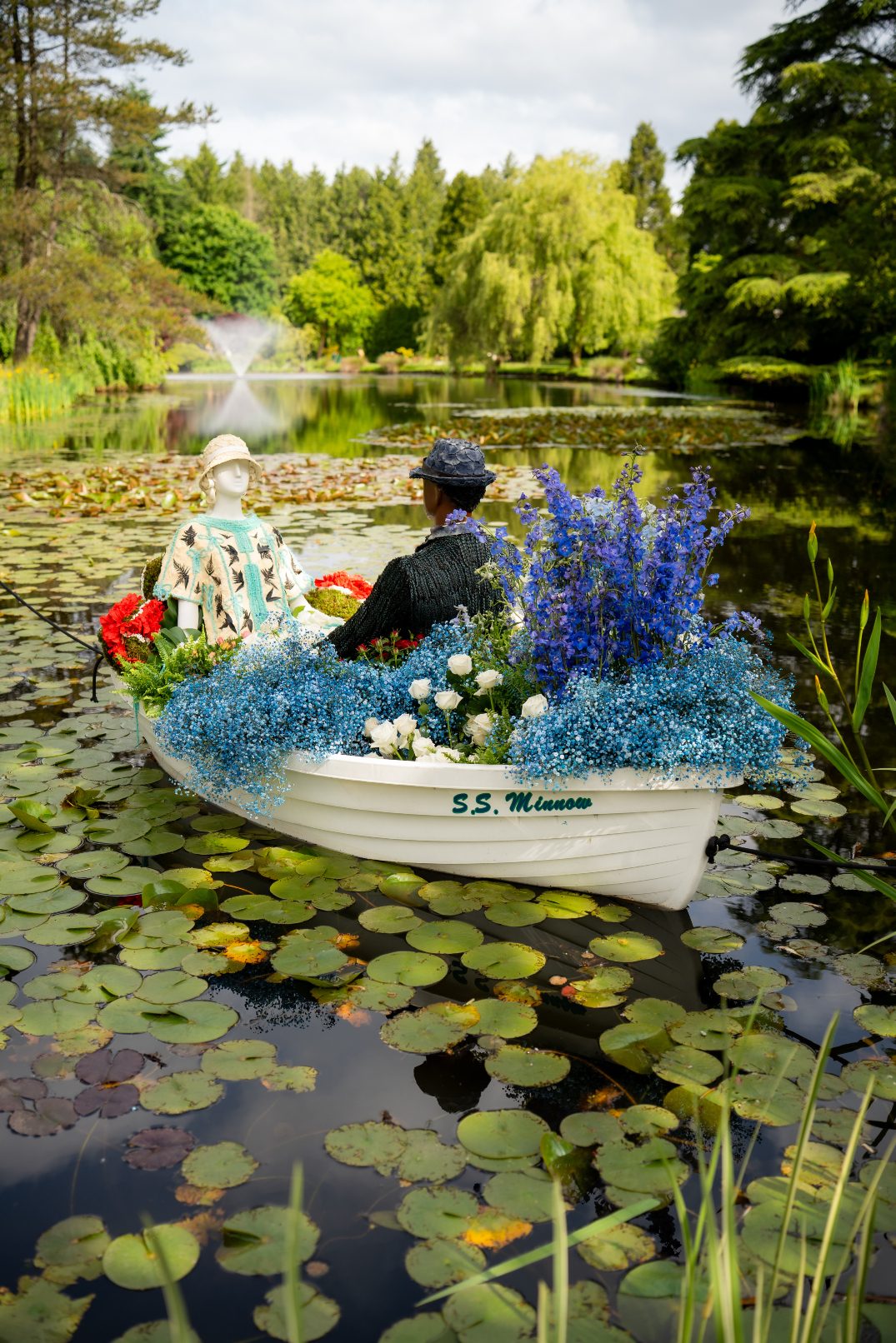 Side view of the S.S. Minnow boat overflowing with blue delphinium, roses, baby's breath and greenery, floating on a pond of lily pads during the Downton Abbey Fleur De Villes event.
