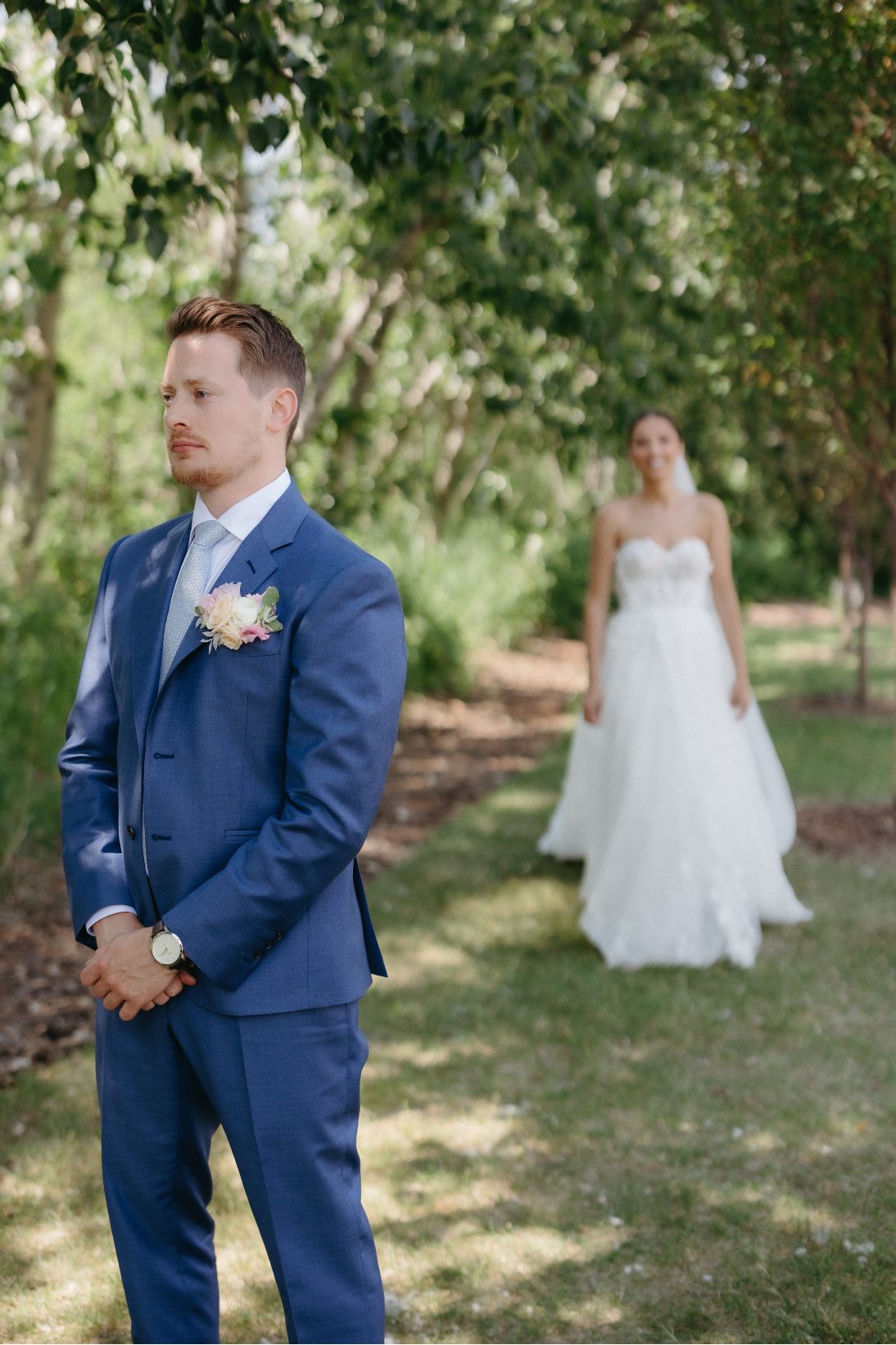 The groom stand facing forward in anticipation with the bride approaching behind him for their first look, both surrounded by dappled light and greenery.
