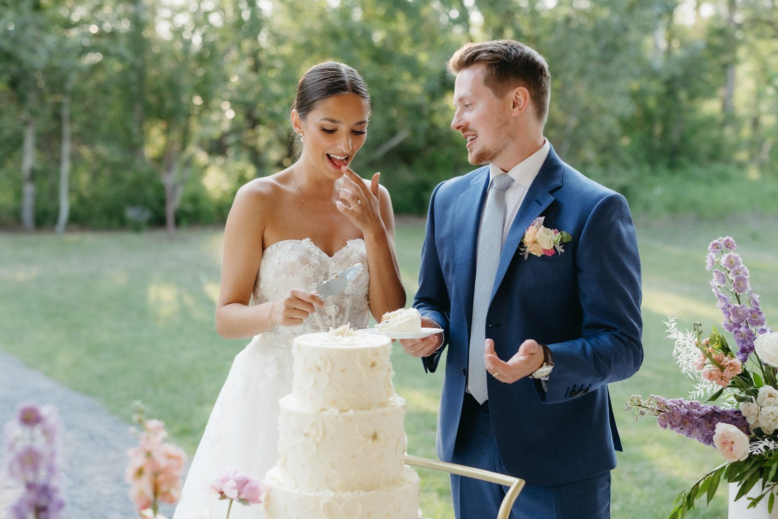The bride licks some icing off of her finger as the groom looks at her, holding a plate with a slice of cake.