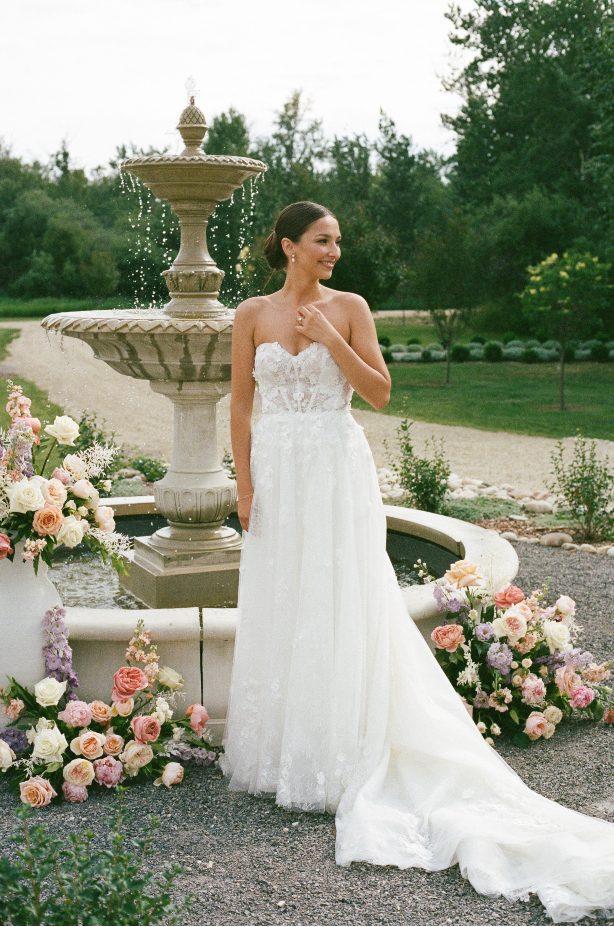The bride stands alone by a grand stone fountain surrounded by pastel floral arrangements, gently touching her collarbone as she looks off and smiles.