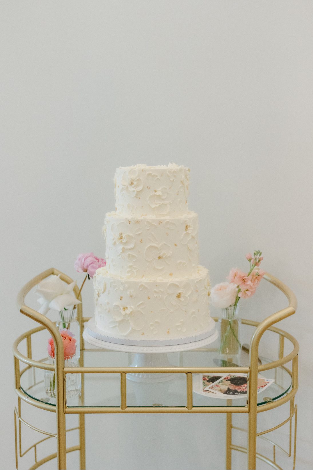 An elegant ivory three tiered wedding cake on a champagne cart accented with a few bud vases with pastel blooms.
