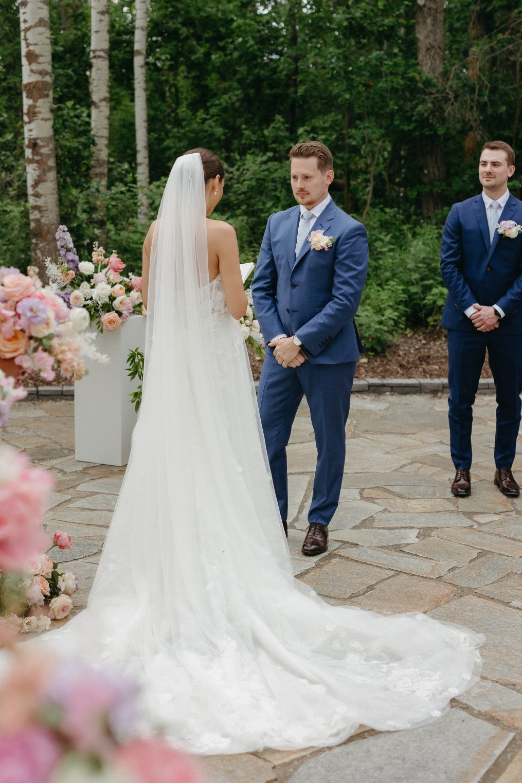 The bride looks stunning with her dress and veil fanned out beautifully behind her while the groom looks at her lovingly during the ceremony.