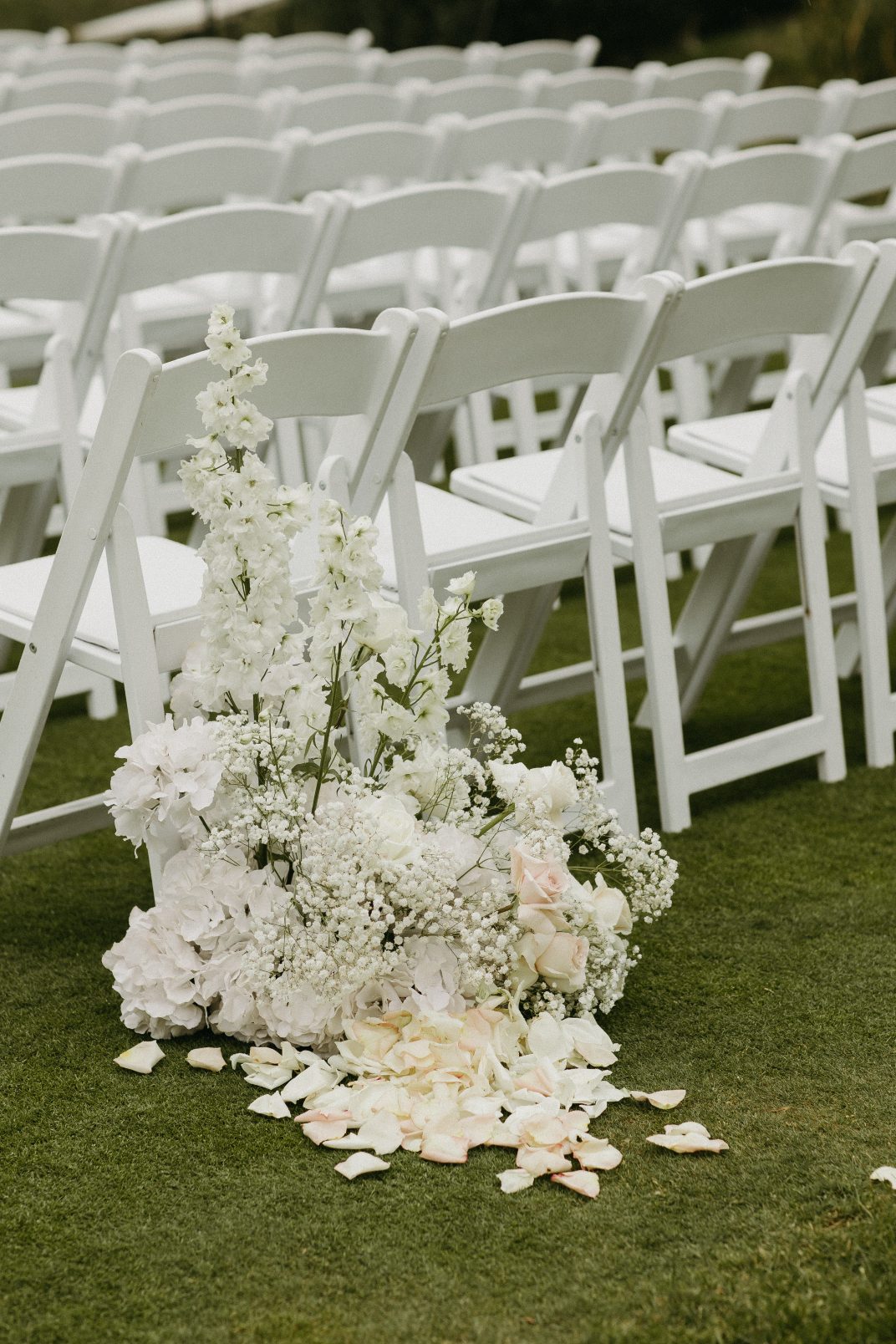 Floral arrangement at the base of a white ceremony chair row, featuring ivory blooms, baby's breath and scattered rose petals on the grass.