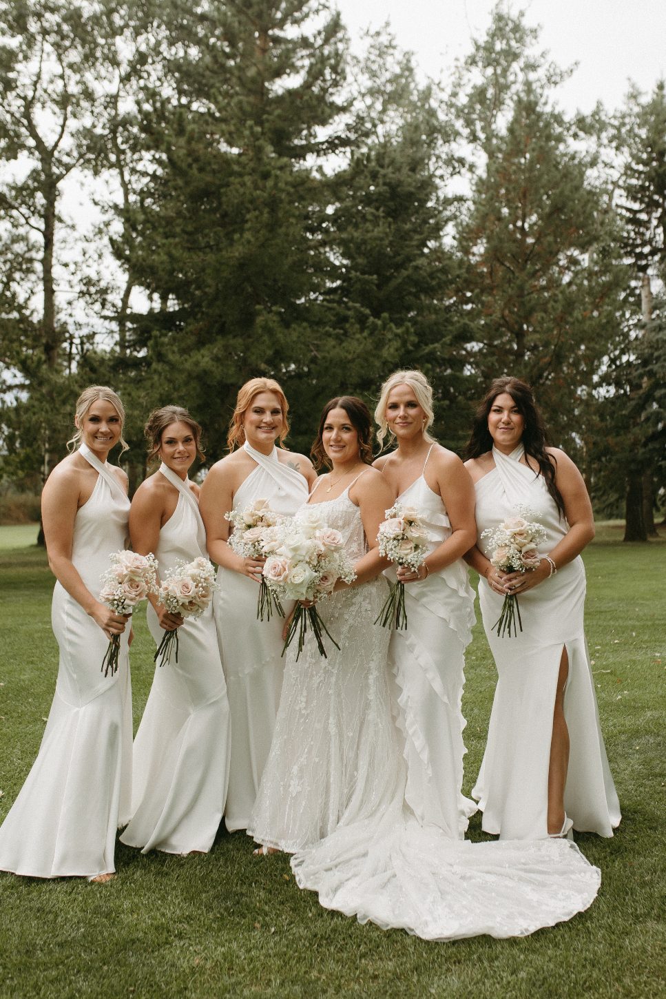 Bride stands centered with bridesmaids in matching white dresses. Each holds a soft pink and white bouquet while smiling outdoors.
