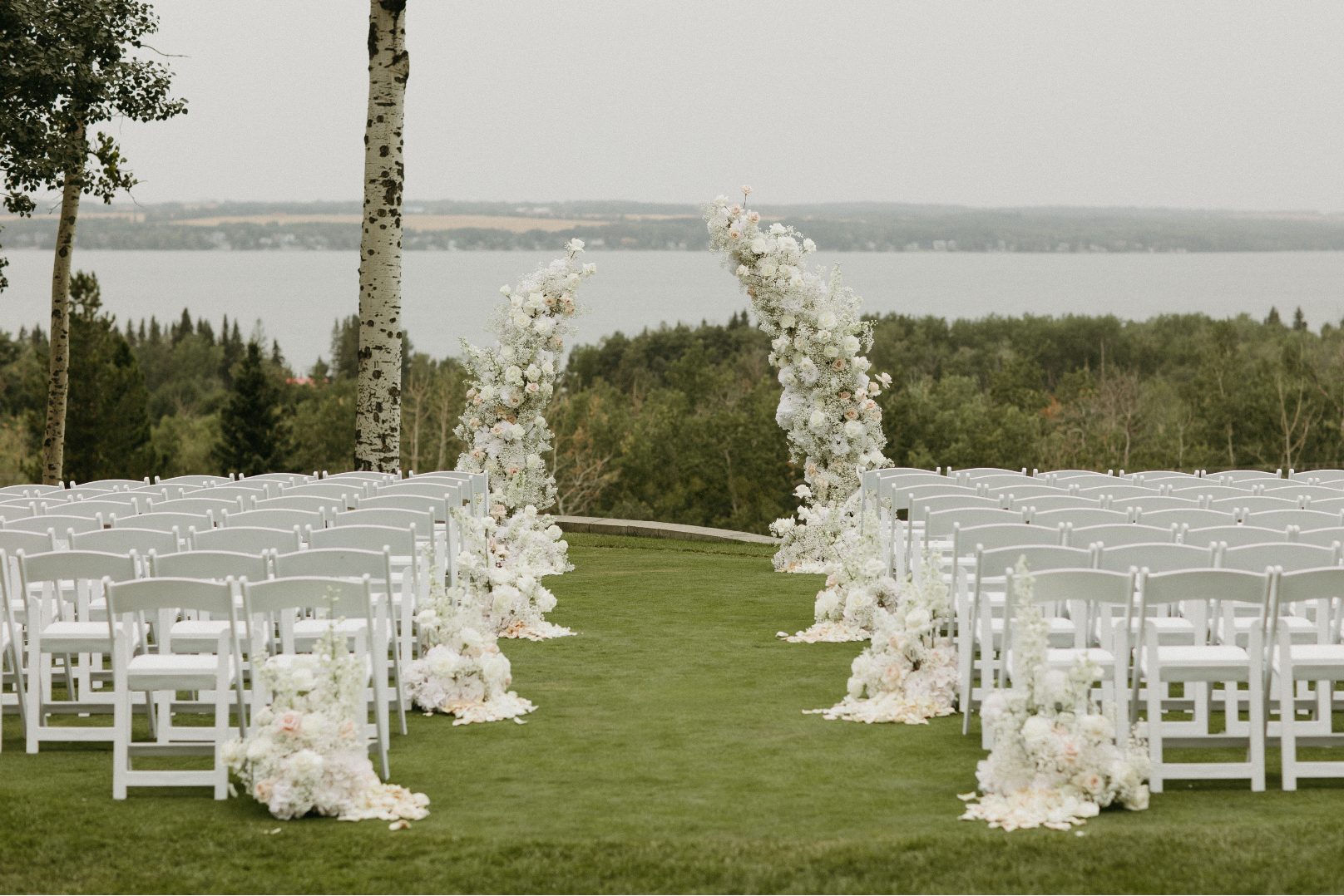 Outdoor ceremony set up with rows of white chairs and a grand floral broken arch framing a scenic lake view, accented with ivory roses and baby's breath.