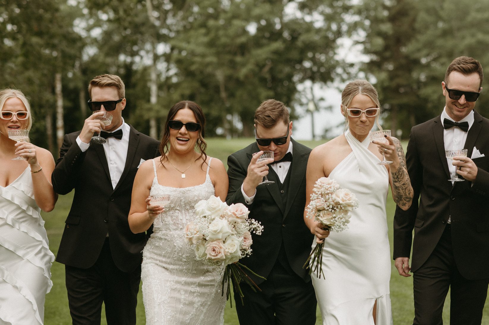Bride and groom walk arm in arm with part of their wedding party, all wearing sunglasses an smiling while holding drinks and bouquets.