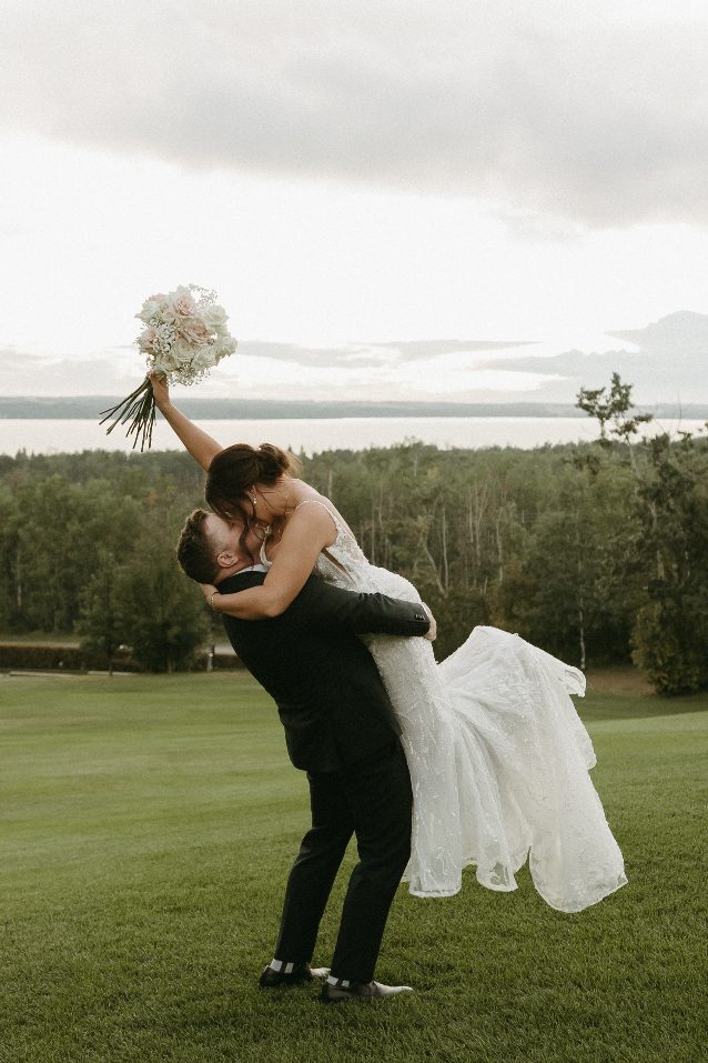 A groom in a black suit lifts his bride in a white lace gown as they kiss outdoors on a grassy hill, with a lake and forested backdrop under a moody, overcast sky.. The bride holds her bouquet triumphantly in the air.