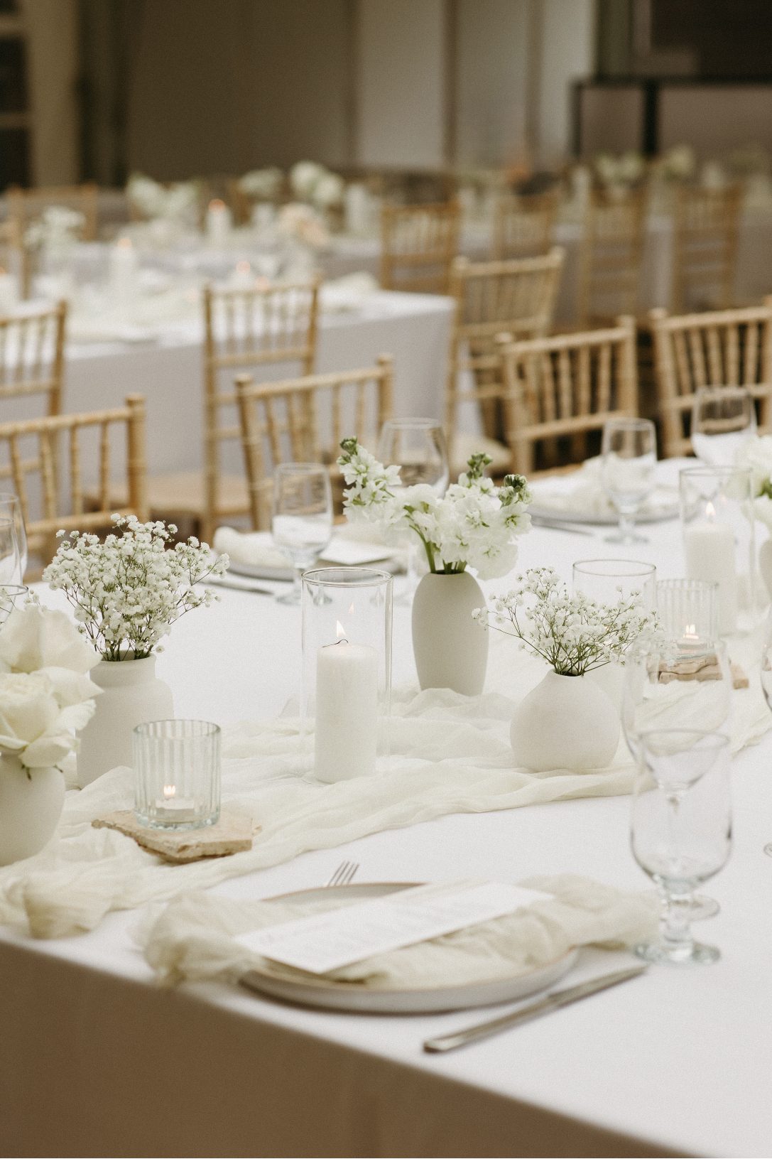 Elegnt white wedding reception tables with soft draping, pale wood chairs, minimalist floral bud vases and romantic candlelight.