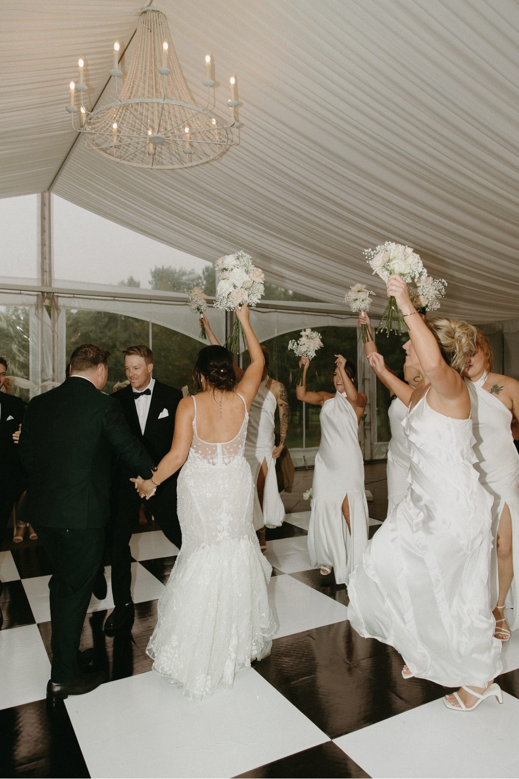 Bride and bridesmaids in wute dancing joyfully with bouquets raised on a black and white checkered dance floor under a tented ceiling.