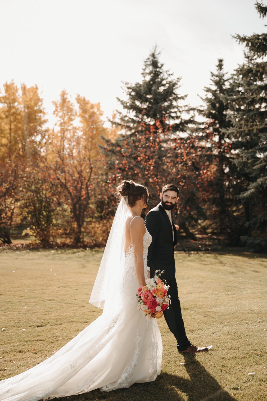 Bride and groom walk outside hand in hand, bride carrying a bright bouquet of coral, pink and peach blooms, surrounded by fall foliage.