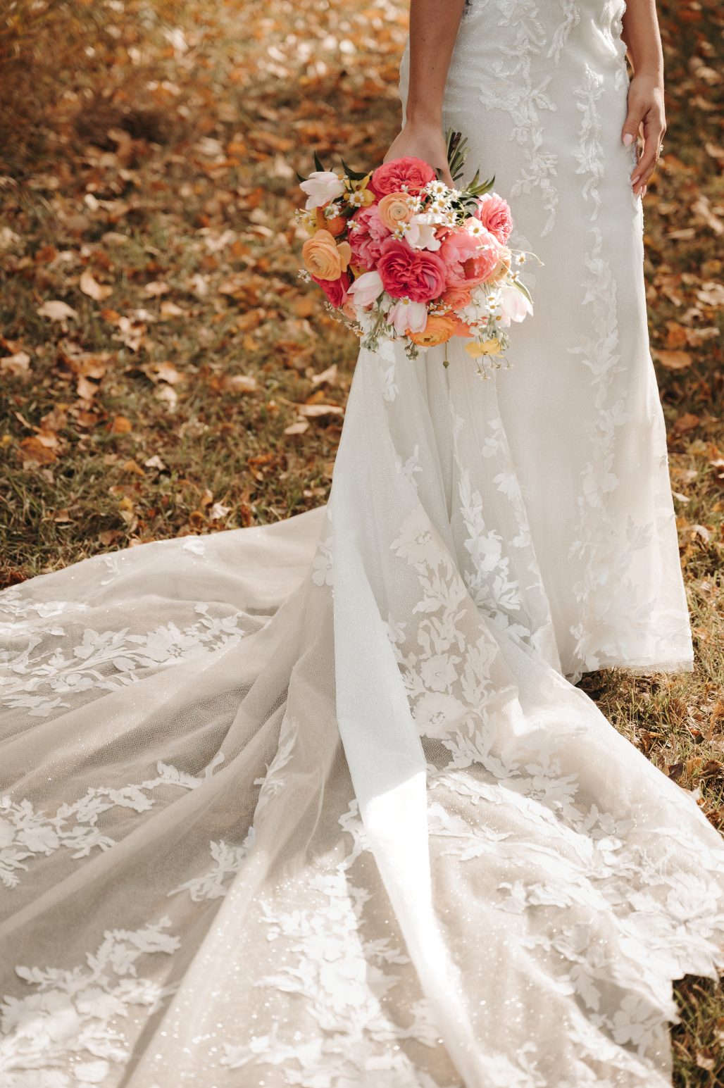 Close up of bride's beautiful lace train spread across fallen autumn leaves, with her vibrant bouquet of coral, pink and peach blooms resting by her side.