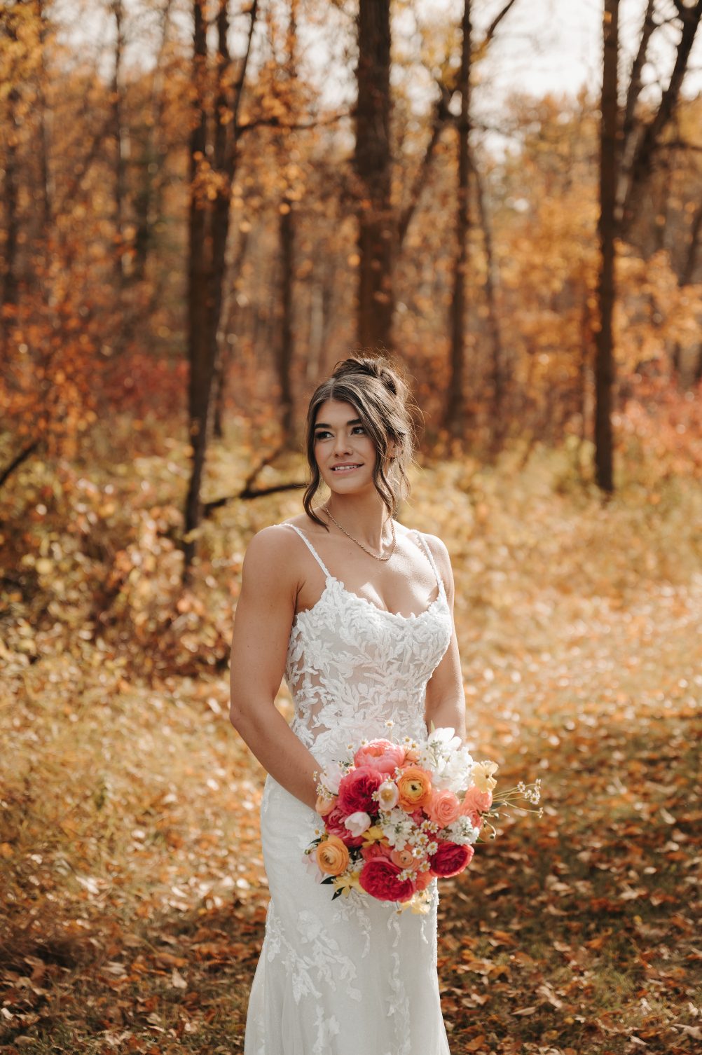 Bride standing among golden autumn leaves holding a bouquet of coral, pin, peach and yellow blooms, wearing a gorgeous fitted gown.