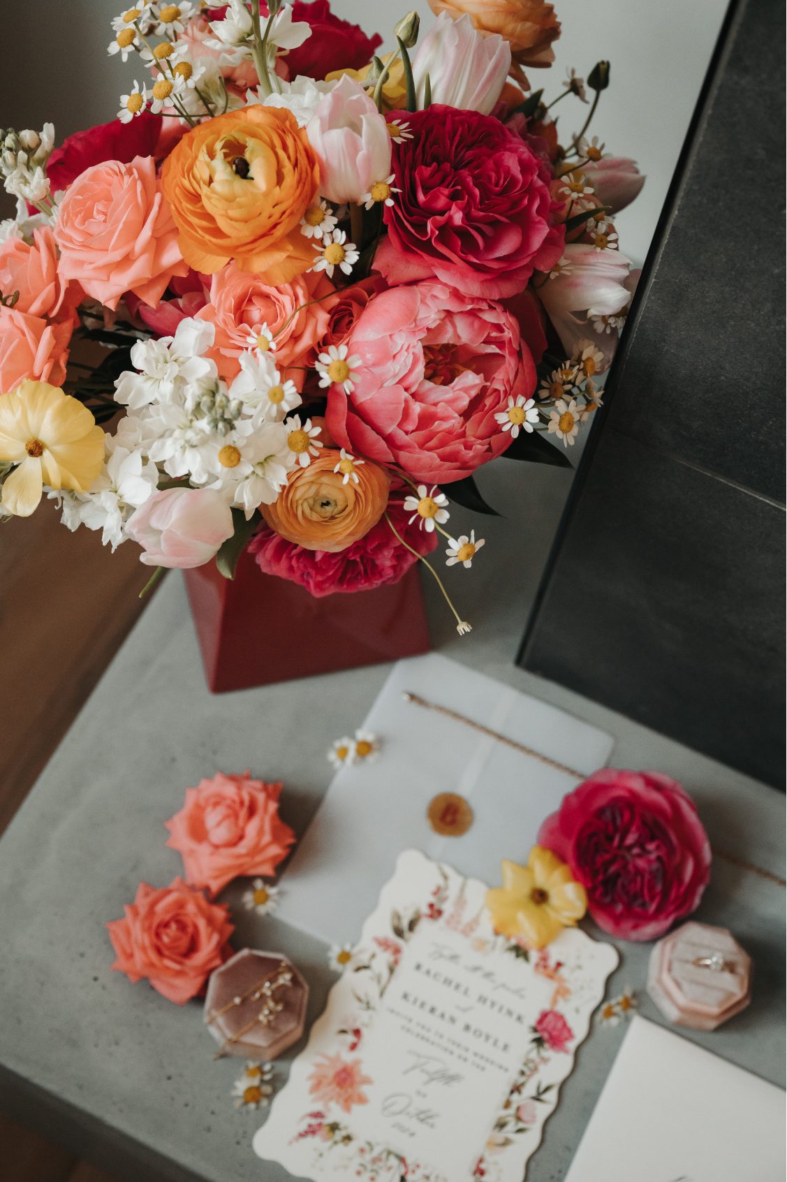Bright wedding bouquet with coral garden roses, peonies, ranunculus, tulips, chamomile and stock beside invitation and rings.
