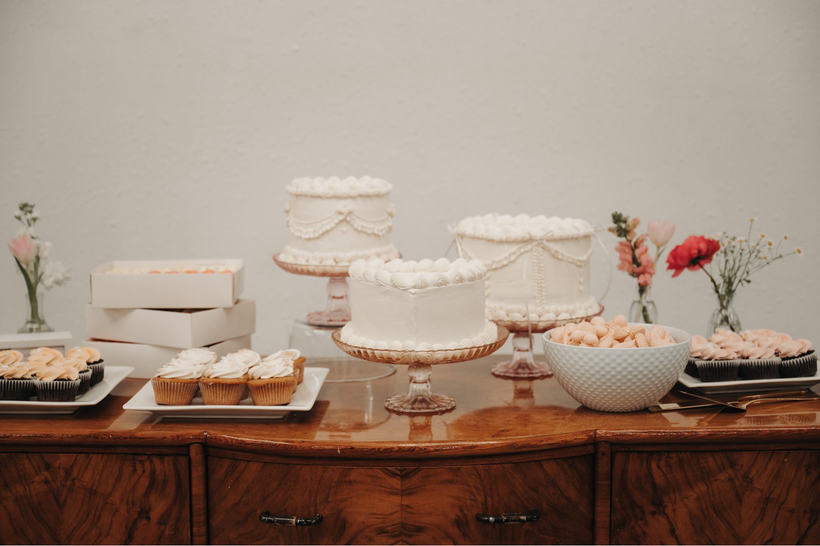 Dessert table with multiple white frosted cakes, cupcakes, and cookies displayed on vintage wooden furniture with simple floral bud vase accents.