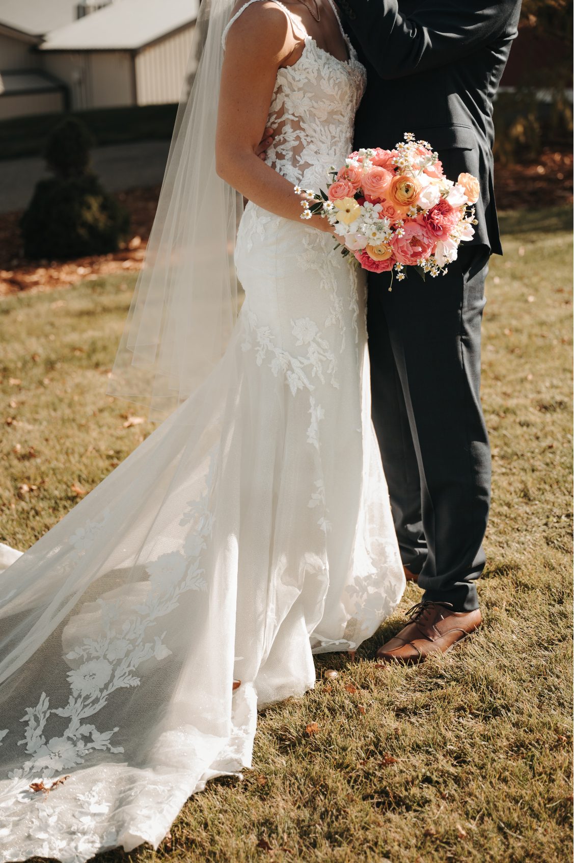 Close up of bride and groom embracing, showcasing the bride's lace gown train and colourful bouquet of coral, peach, pink and yellow flowers.