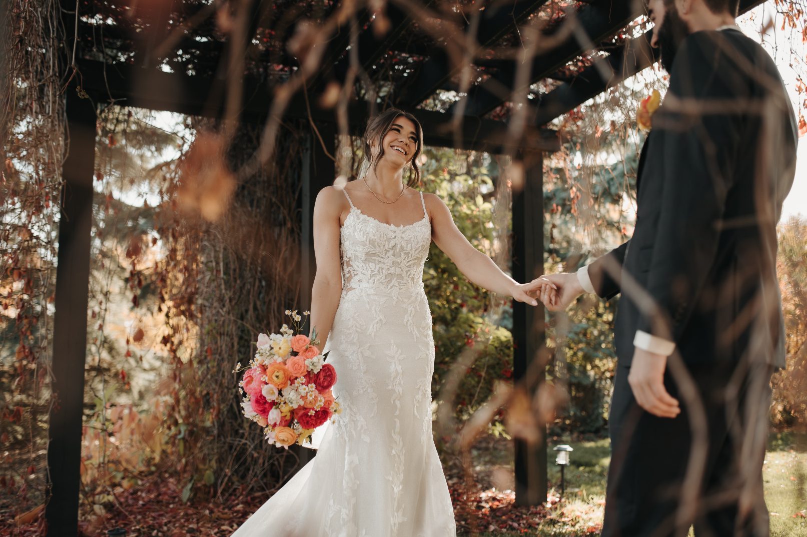Bride smiling vibrantly as she holds a beautiful bouquet of coral, peach, pink and yellow, reaching for the groom's hand under a wooden arbor.