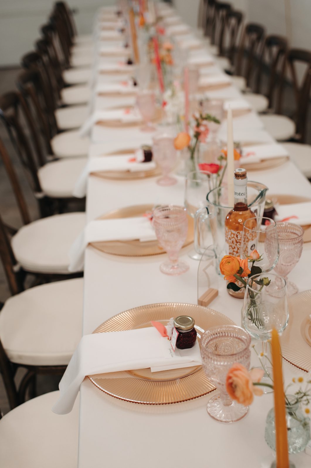 Long wedding reception tables set with blush goblets, gold chargers, taper candles and small floral bud vases down the center.