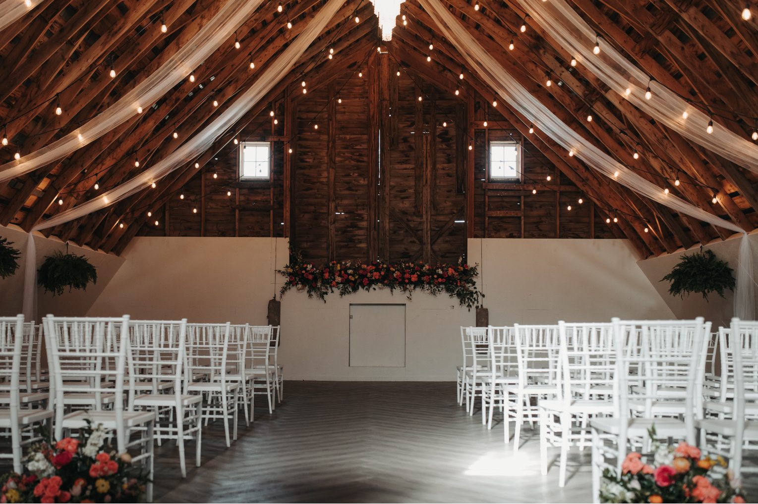 Barn wedding ceremony setup with white charis, wooden beams draped in fabric, and a bold floral altar installation under glower string lights.
