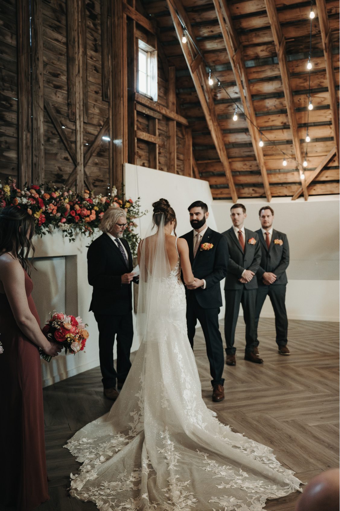 Bride and groom holding hands at the altar with officiant and wedding party, with a vibrant floral backdrop.