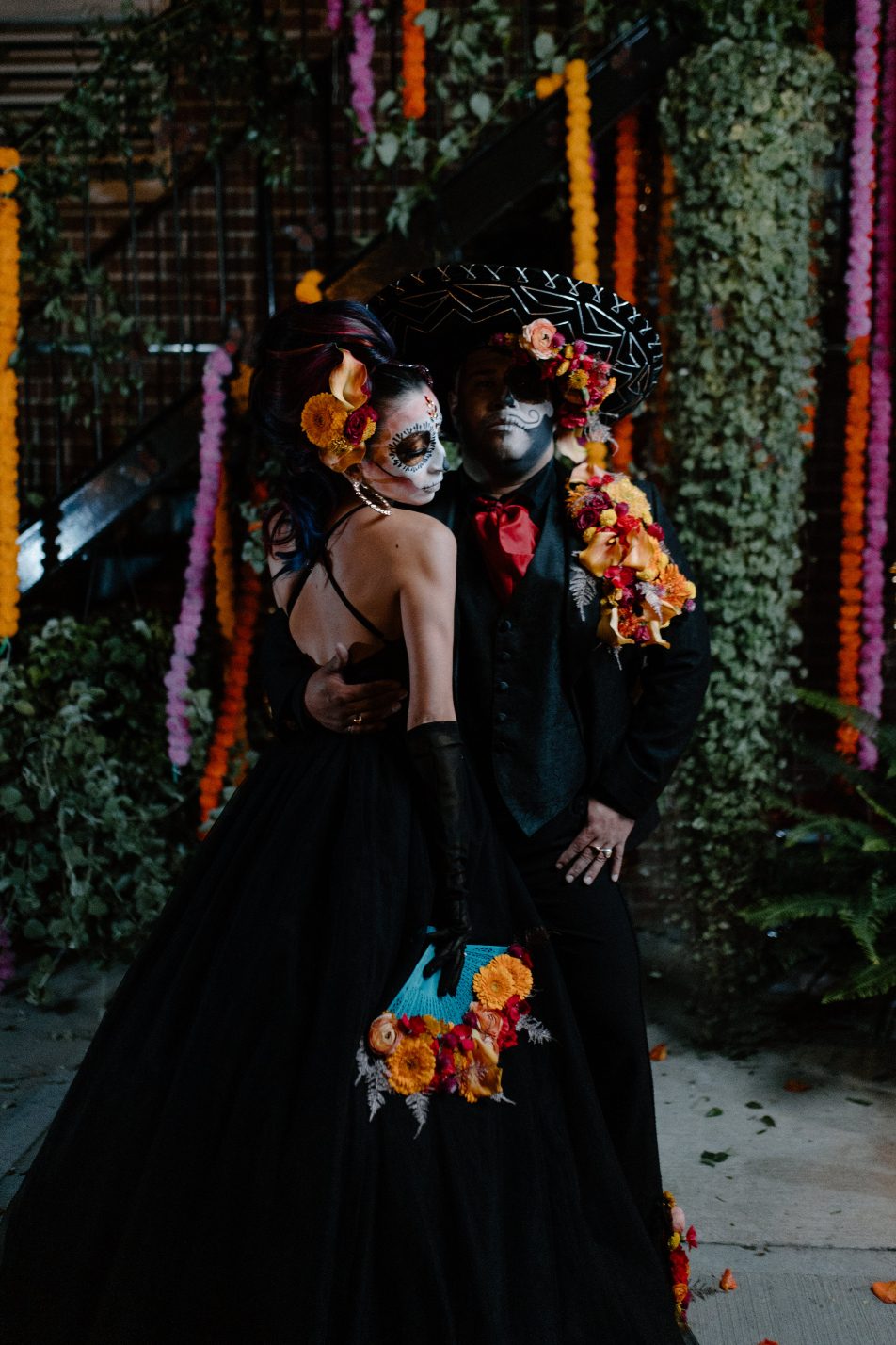 Couple dressed in black with bold floral accents share a romantic embrace in front of vibrant marigold garlands.