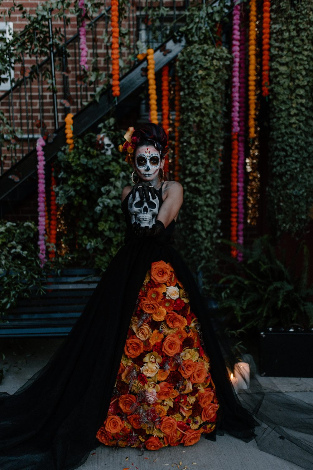 Model in floral adorned black gown holds skull, surrounded by lush foliage and colourful hanging marigold strands.