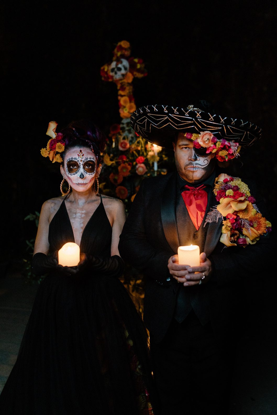 Couple in black attire with bold floral accents stand side by side each holding a lit pillar candle and staring at the camera.