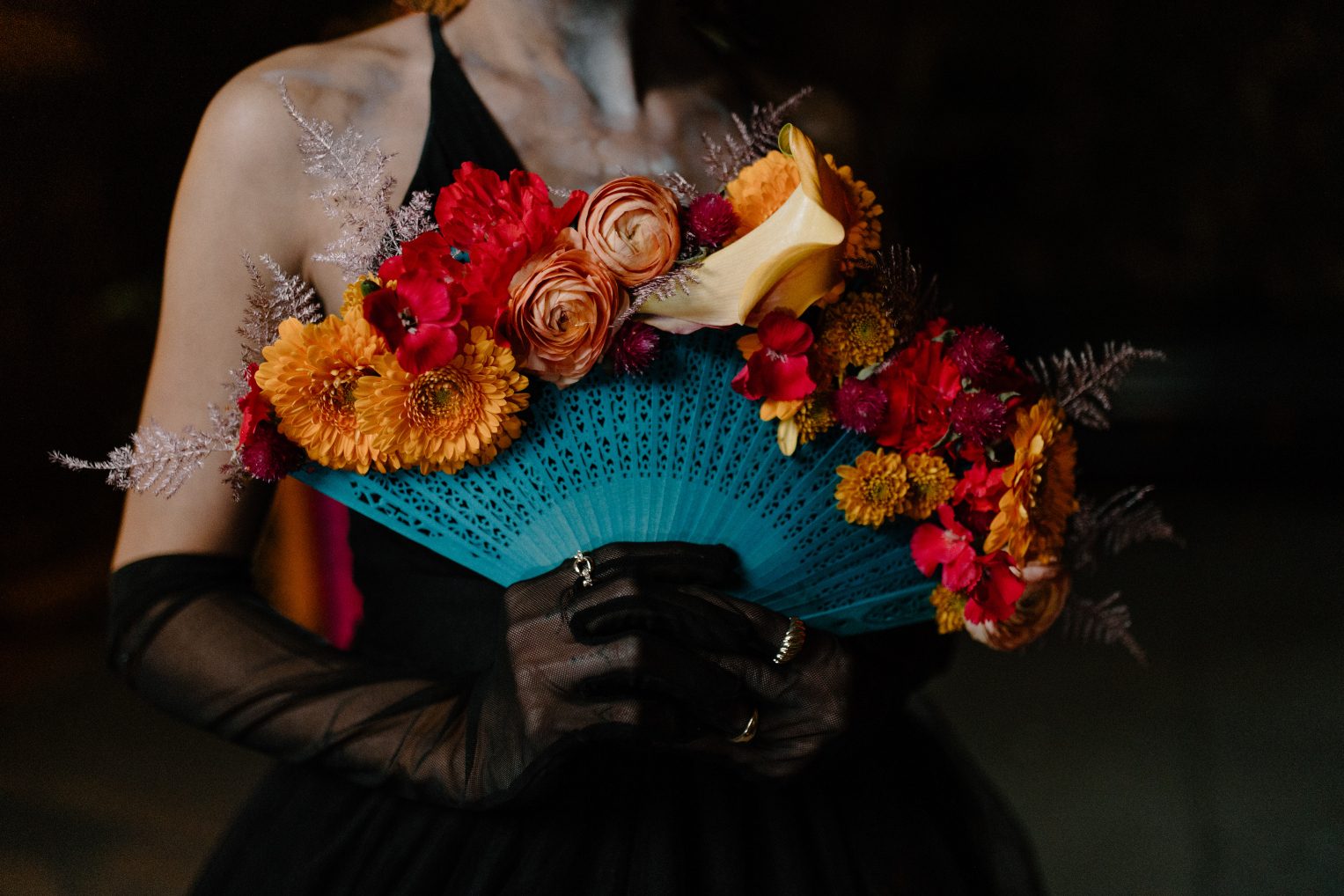 Close up of floral hand fan featuring orange gerbera daisies, ranunculus, calla lilies and red accent blooms.