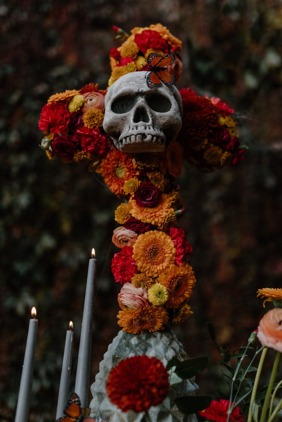 Floral cross featuring a skull centrepiece, marigolds, ranunculus and monarch butterfly against dark foliage backdrop.