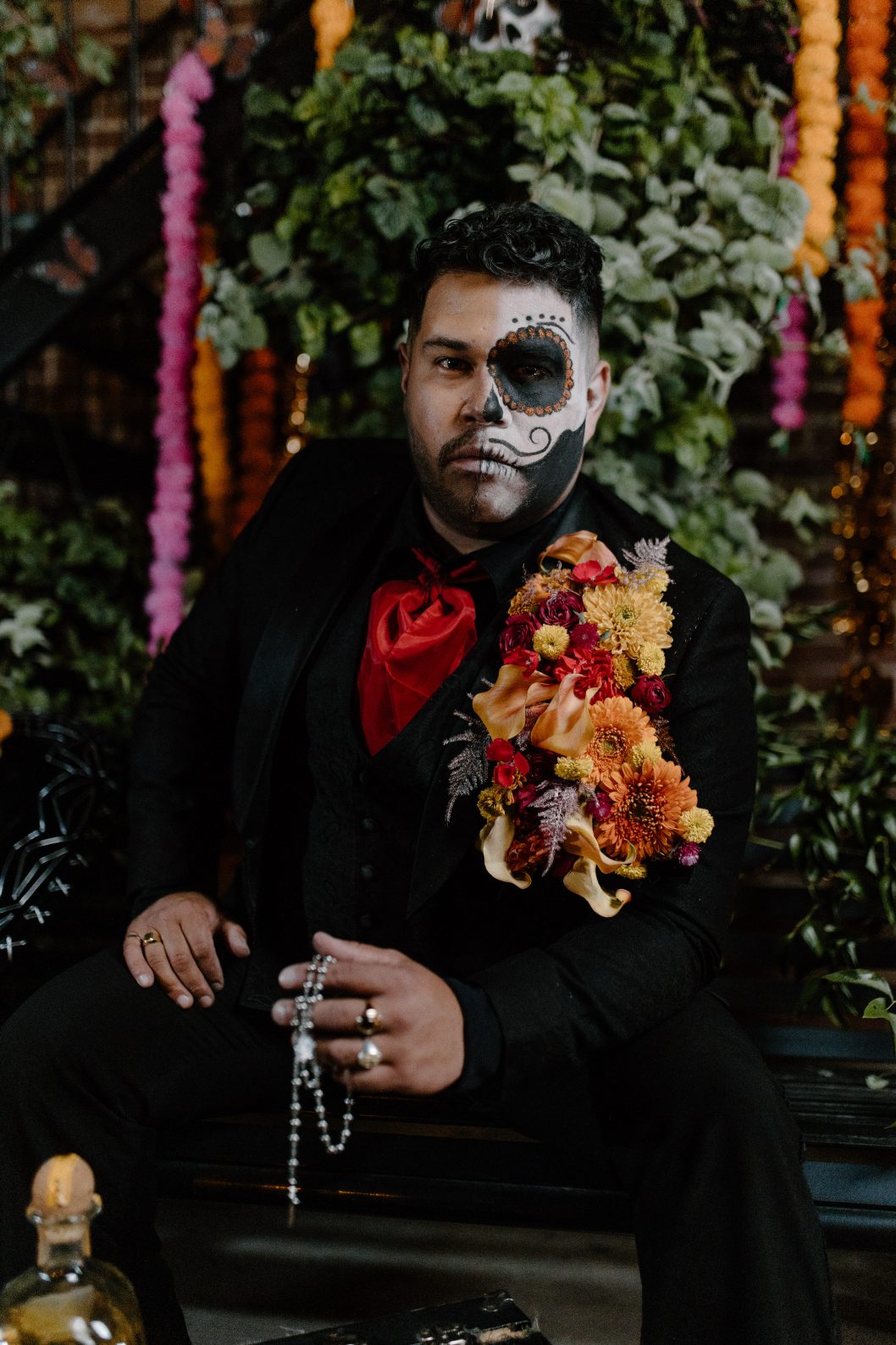 Groom seated in a black suit with floral shoulder piece and rosary, surrounded by hanging garlands and foliage.