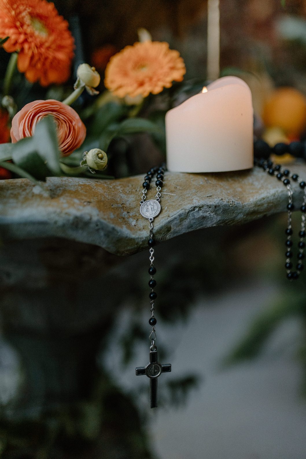 Close up of the altar with a candle and a rosary draped over the stone, surrounded by vivid orange blooms and lush greens.