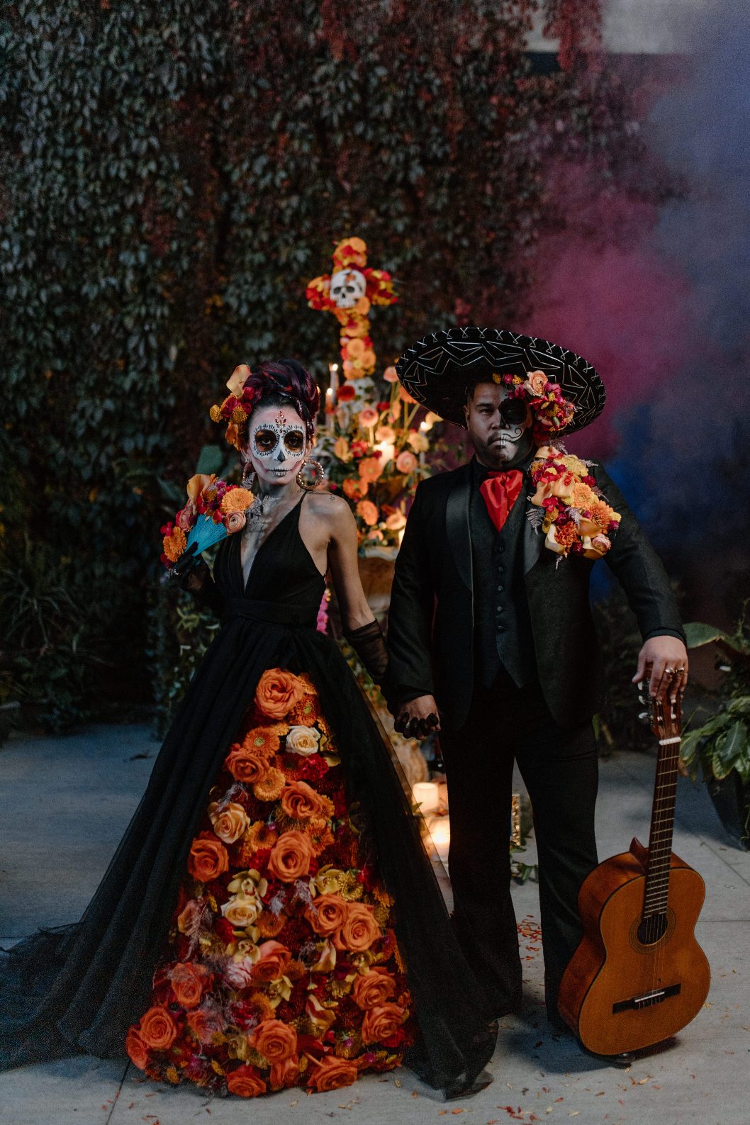Couple in elaborate floral attire stands before altar with floral skull cross and soft pink and blue smoke in the background.