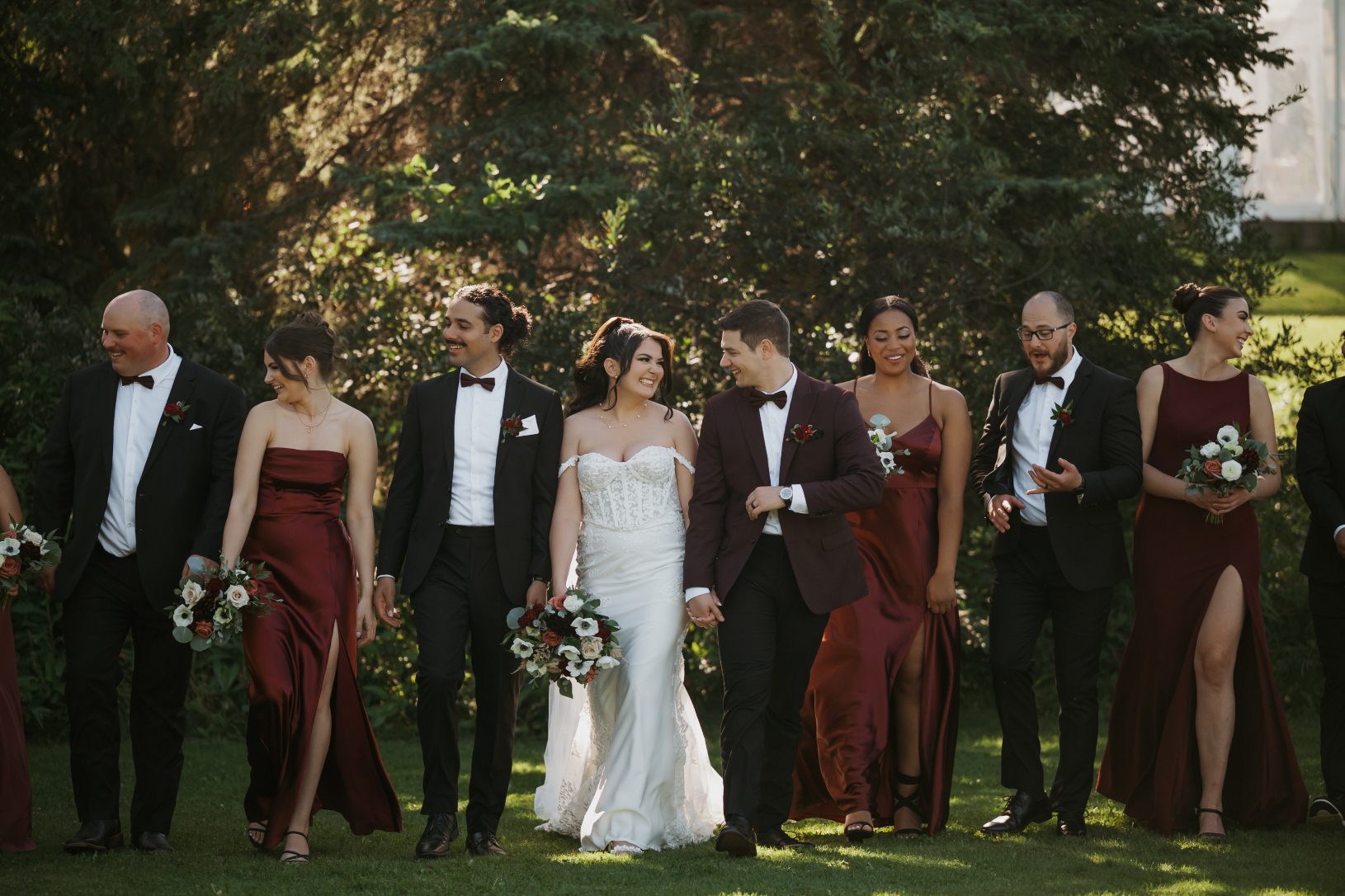 Bride and groom walking with their wedding party on a sunlit lawn, with bridesmaids in burgundy dresses and groomsmen in black suits.