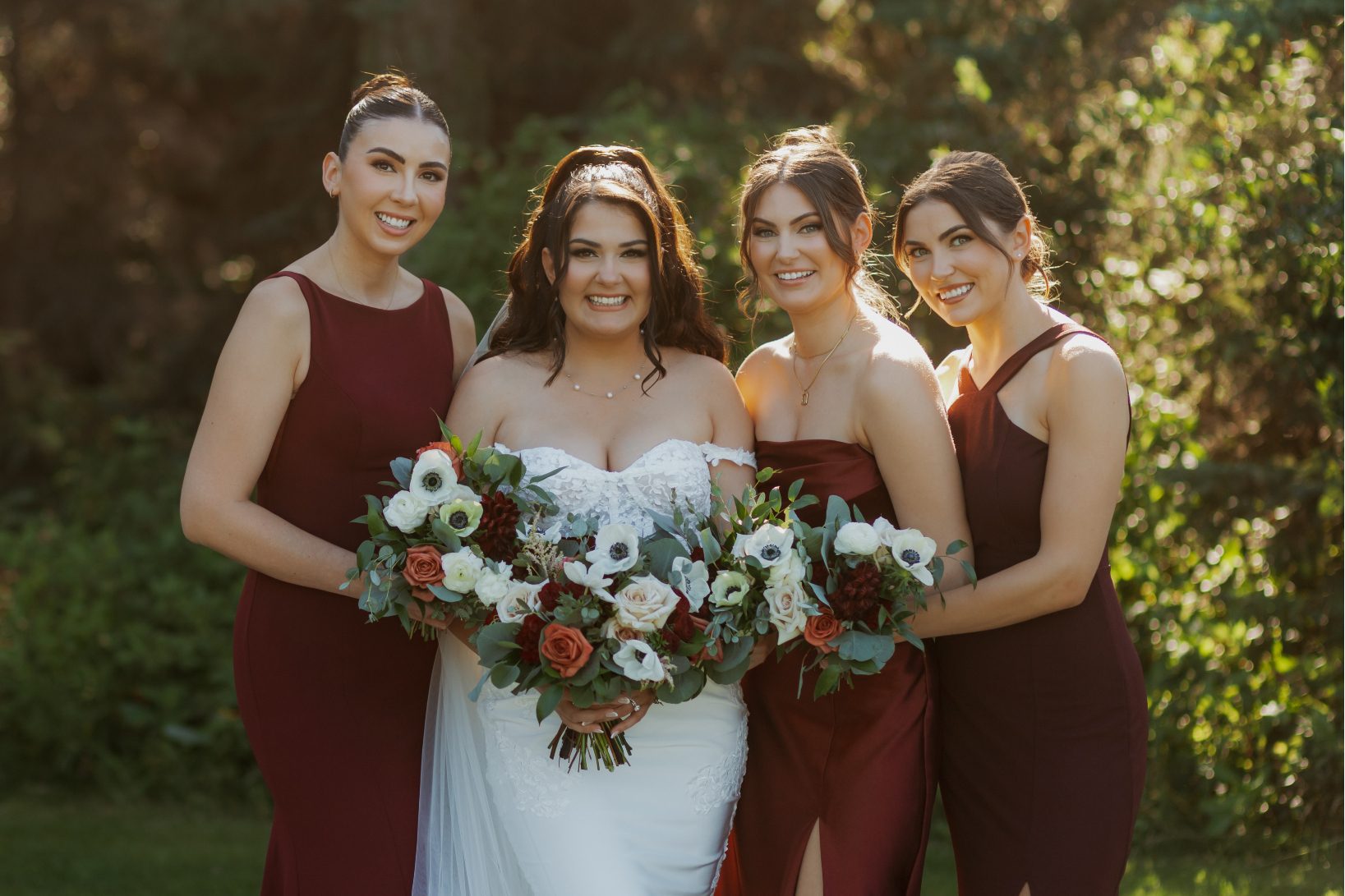 Bride with bridesmaids standing together in deep burgundy dresses, each holding a bouquet with white anemones, roses and greenery.