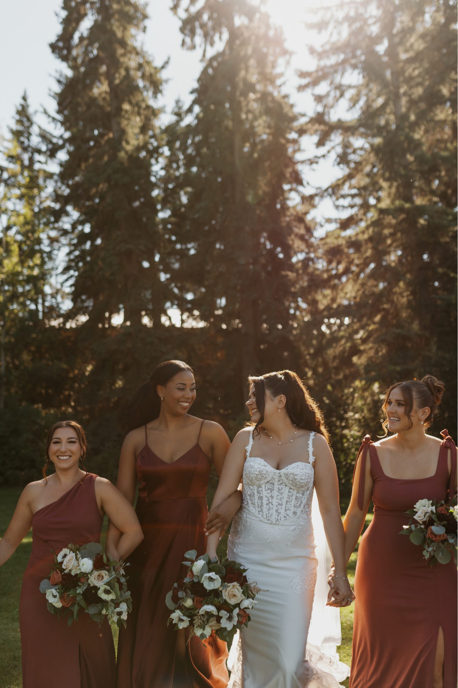 Bride walks arm in arm with her bridesmaids in mixed burgundy gowns, holding romantic white, blush and burgundy bouquets.