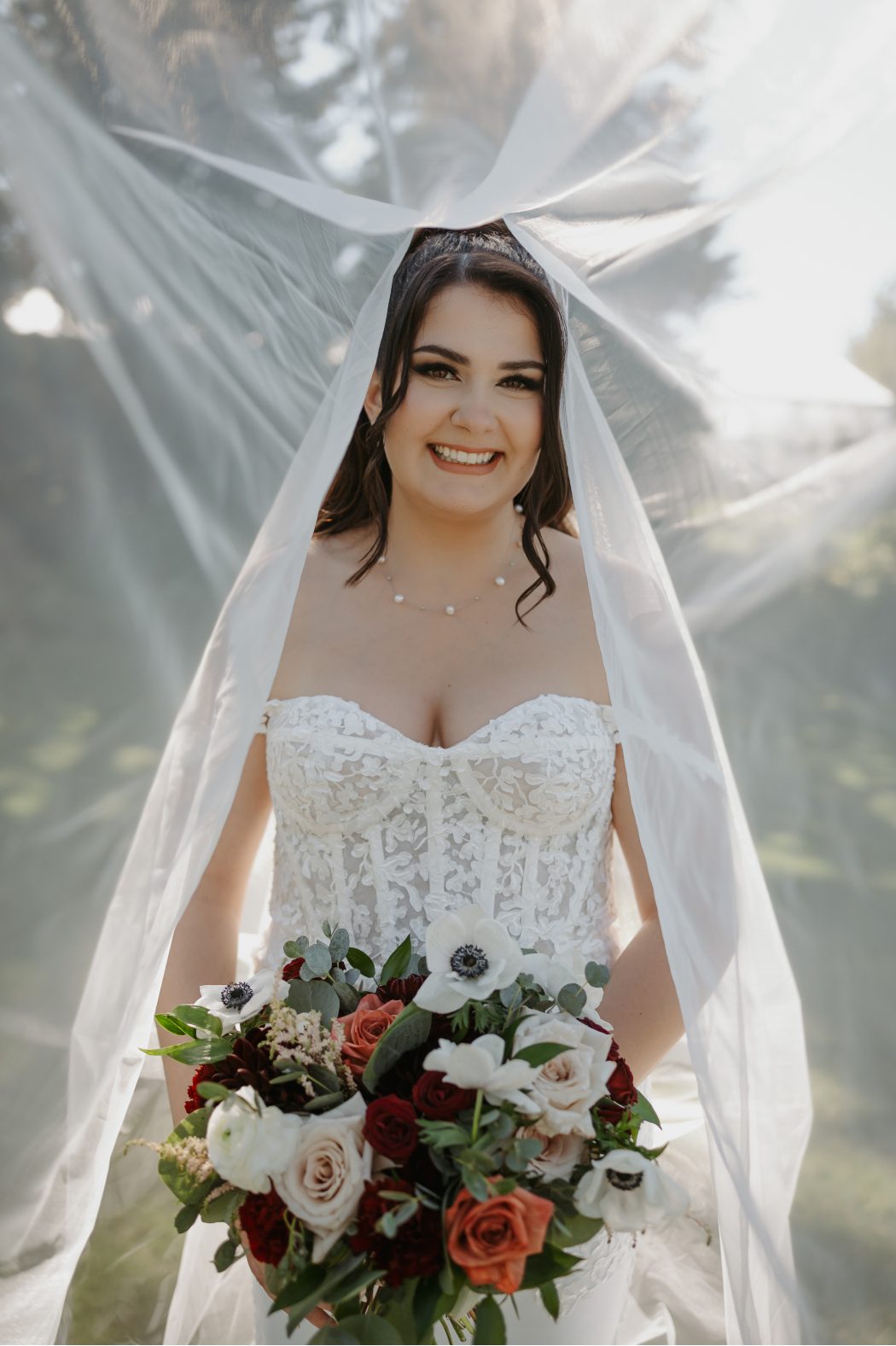 Bride smiling under a flowing veil, wearing a lace corset gown and holding a bouquet of anemones, roses, dahlias and eucalyptus.