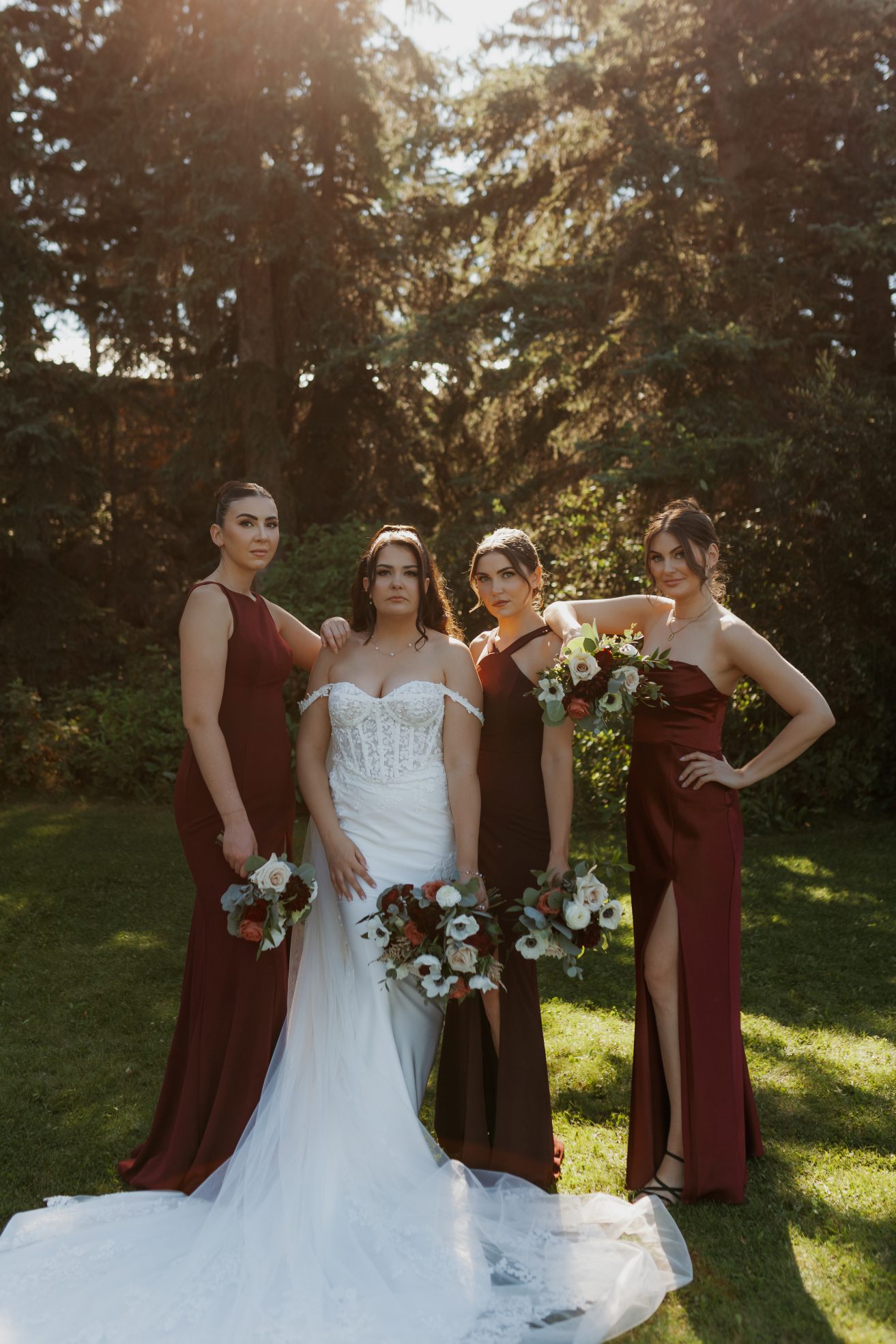 Bride standing with three bridesmaids in fitted burgundy dresses, each holding bouquets and striking a serious pose.