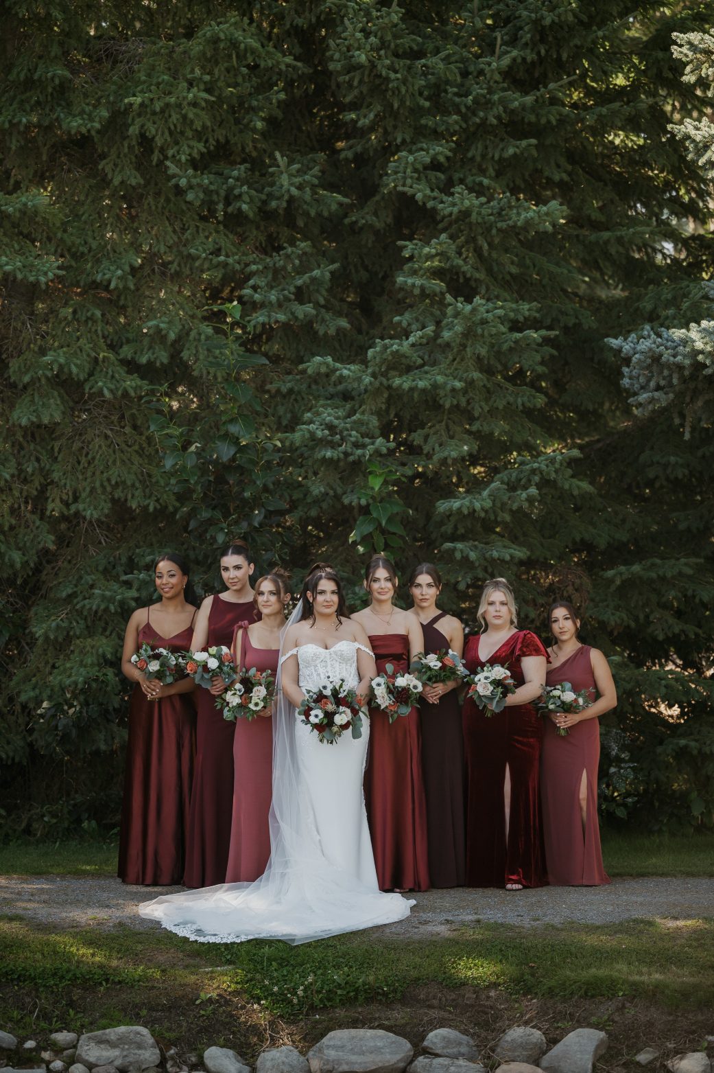 Bride surrounded by bridesmaids wearing assorted burgundy gowns, all holding coordinatd bouquets with roses, anemones, dahlias and eucalyptus.