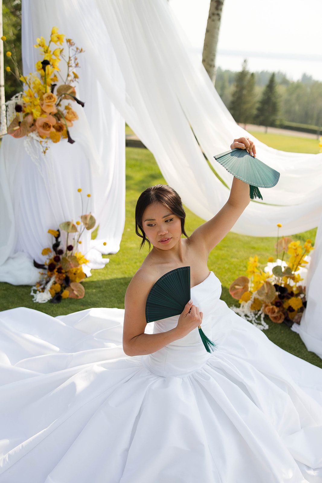The bride sitting in the grass at the ceremony location posing with two teal toned fans.