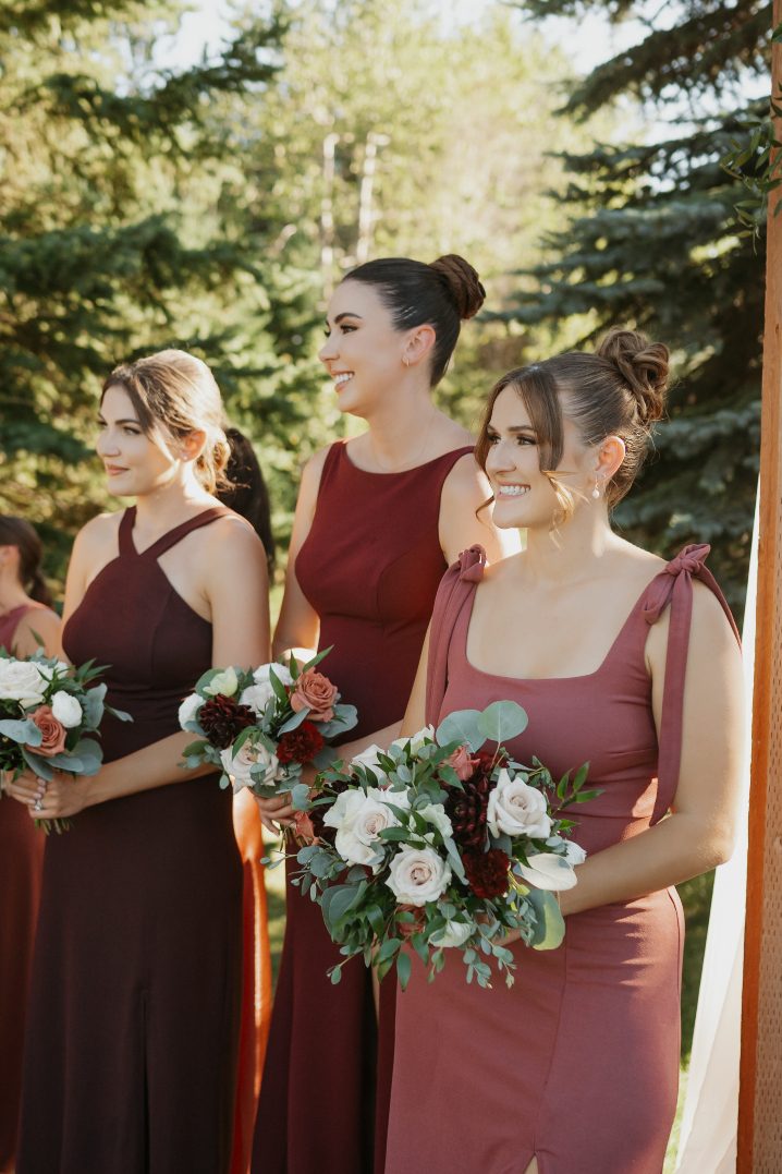 Bridesmaids in burgundy dresses stand holding bouquets, smiling softly as sunlight filters through the trees during the ceremony.