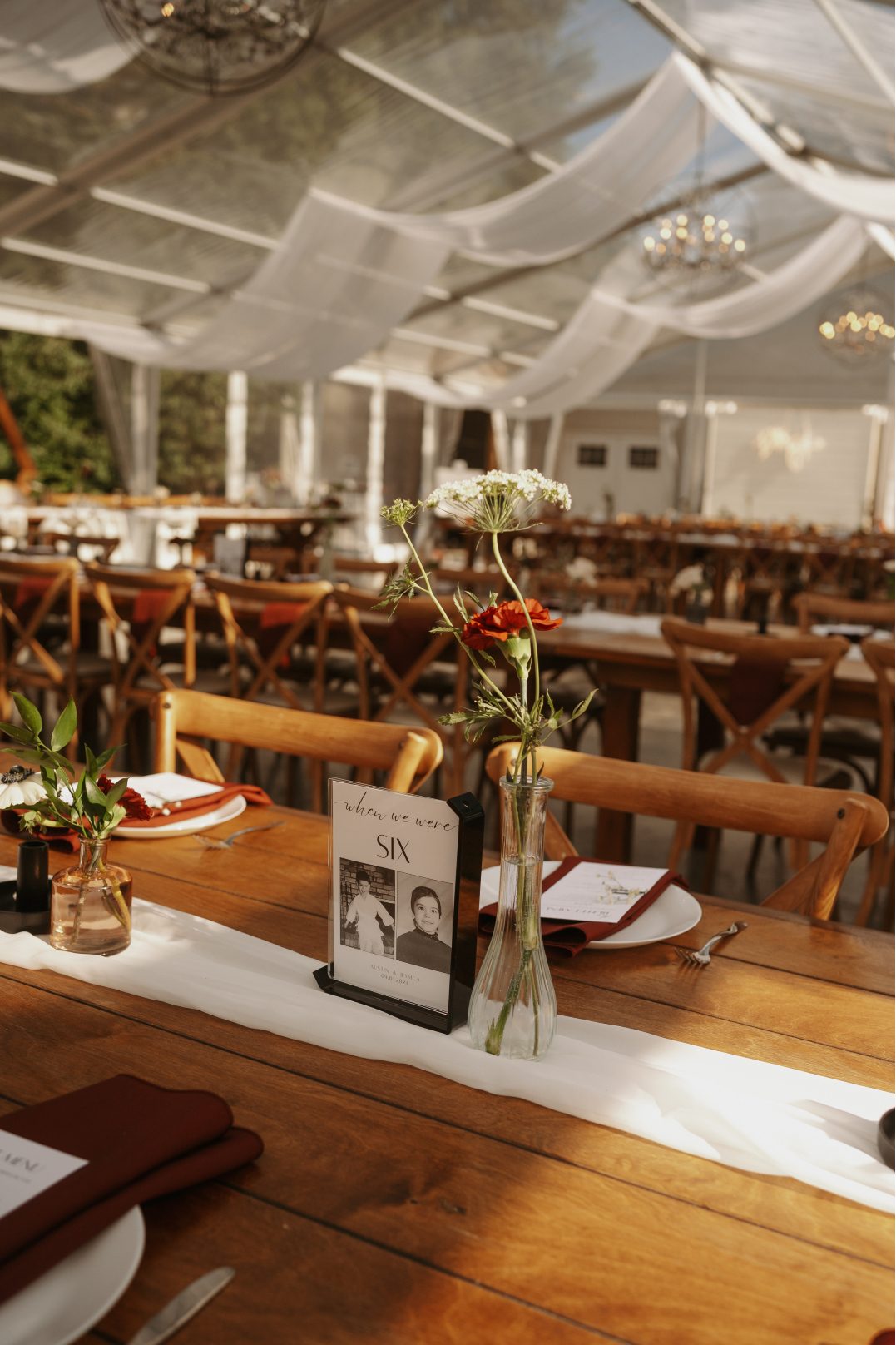 Wedding table centrepiece featuring a tall clear bud vase with a red carnation and Queen Anne's lace besides a table number sign and burgundy place setting,