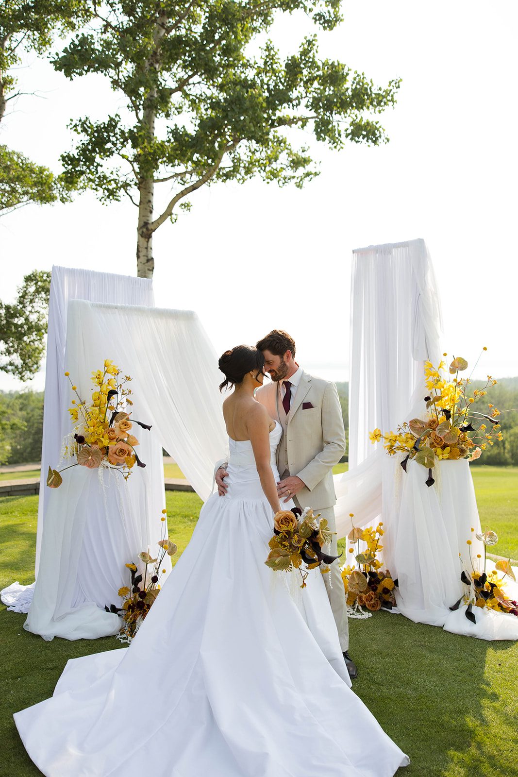 The bride and groom share an embrace about to kiss in the ceremony location, surrounded by bold yellow arrangements and romantic panels of white draping.