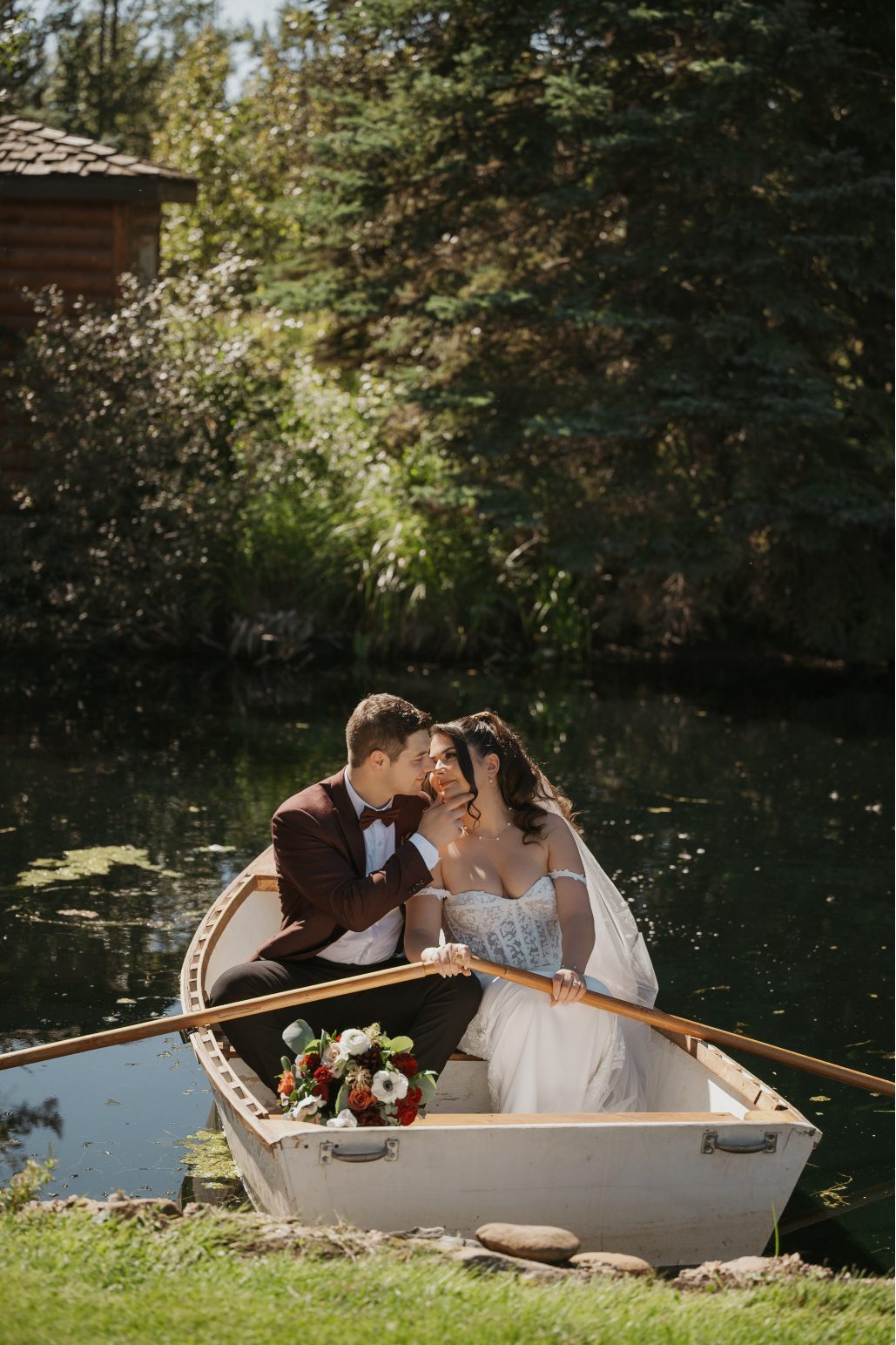 Bride and groom share a kiss while sitting in a wooden rowboat on the pond, with a bouquet resting near the oar.