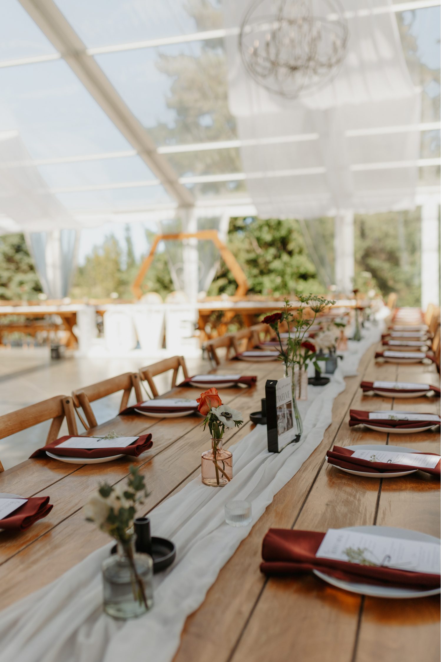 Reception table decorated with a white runner, scattered bud vases, and burgundy napkins, with a clear tent and wooden hexagon backdrop visible in the distance.