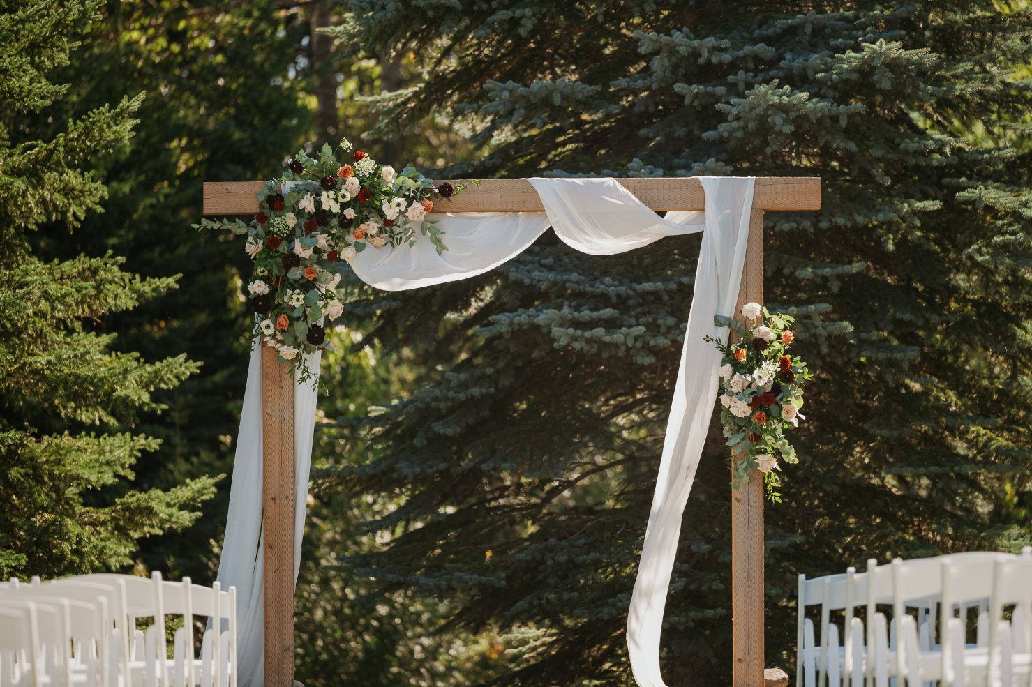 Wooden wedding arch draped with flowing white fabric and decorated with lush floral arrangements in burgundy, blush and white set among tall evergreens.