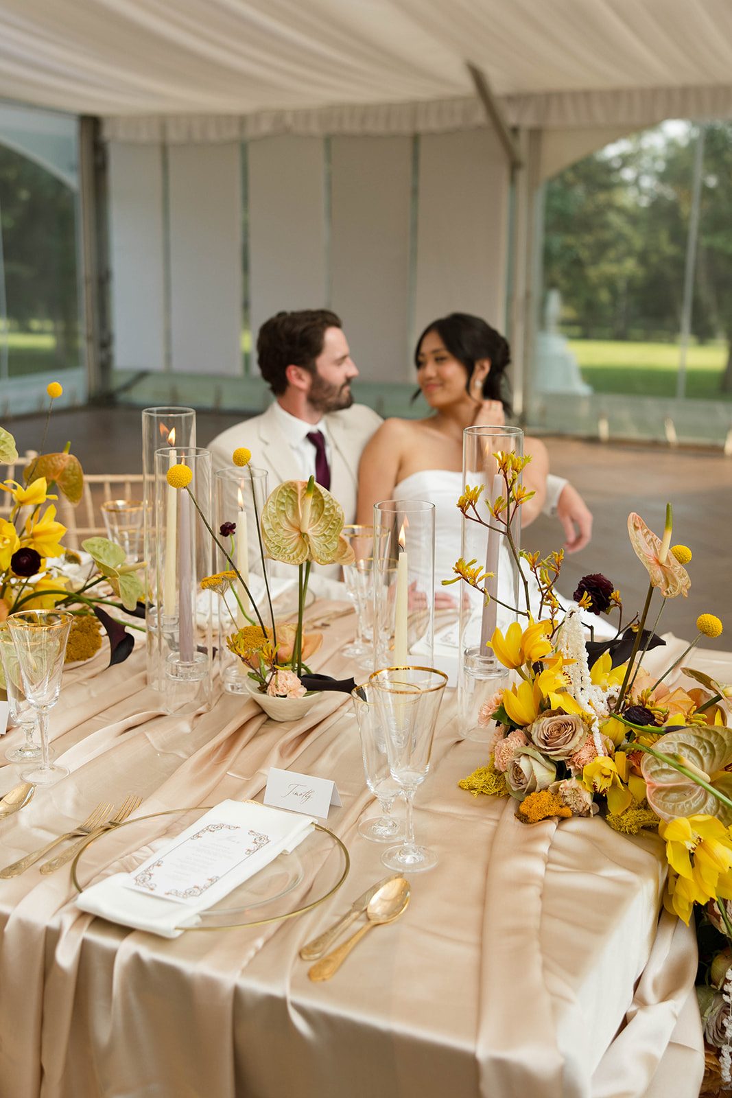 Elegant champagne draped wedding tablescape featuring yellow orchids, anthurium and tapered candles, with a bride and groom smiling in the background.