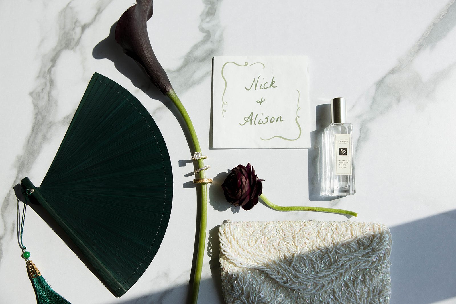 A flat lay shot showcasing the teal fan, brides beaded clutch, ranunculus with rings on the stem of a calla lily.